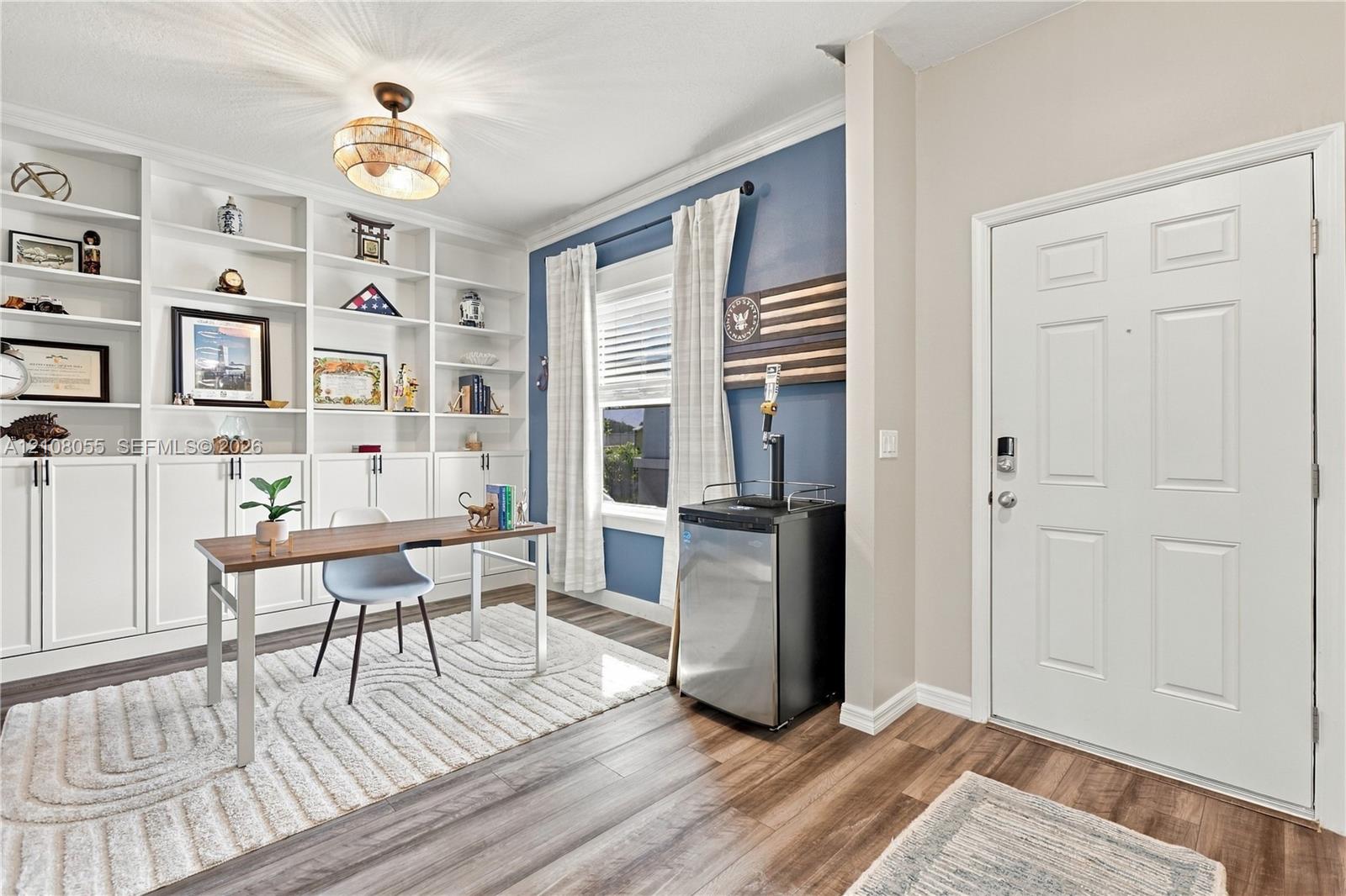 Built-in floor-to-ceiling white shelving with lower cabinetry, wood-finish flooring, multi-pane window with trim, decorative ceiling-mounted fixture, and a six-panel entry door