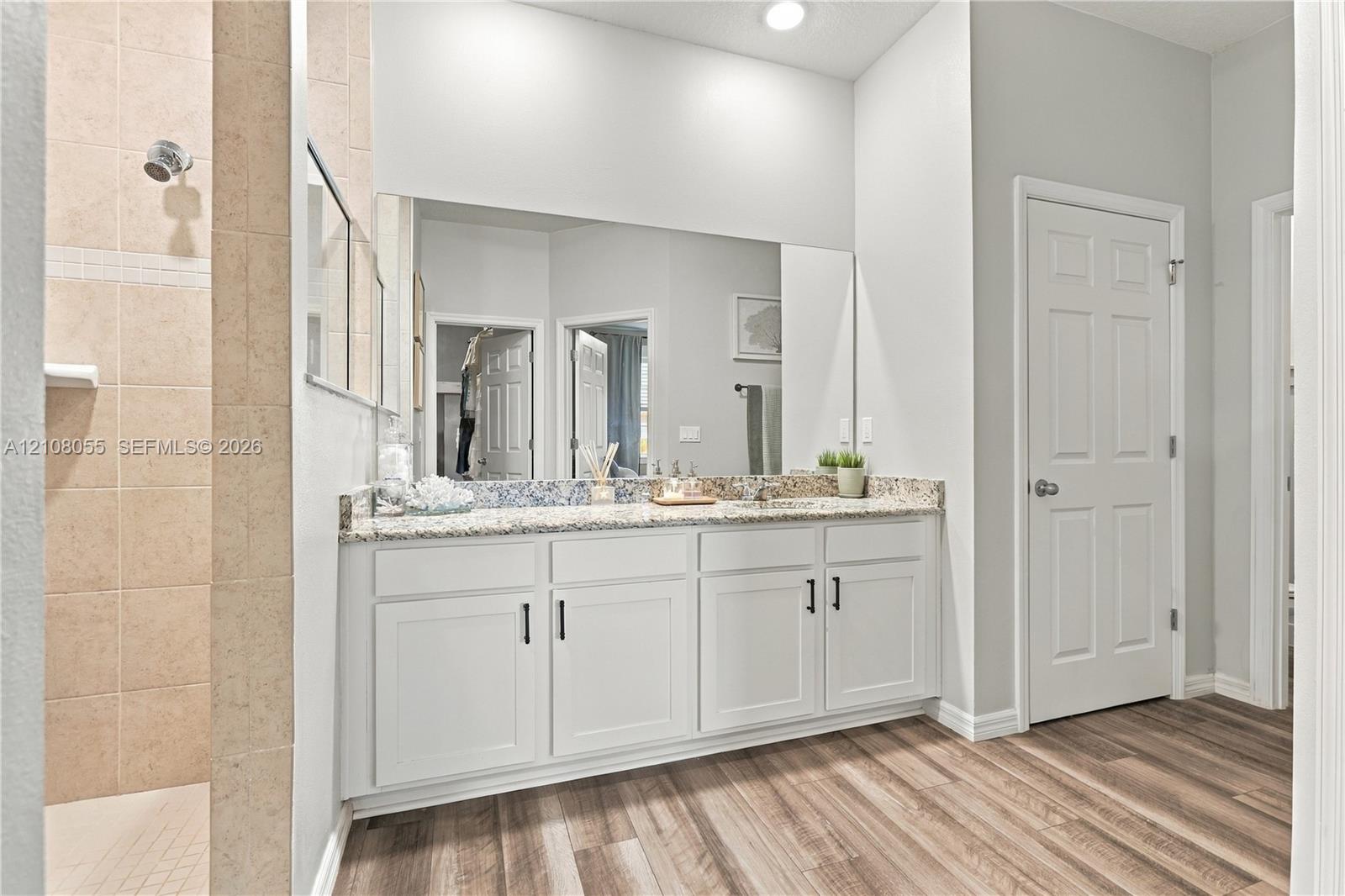 Bathroom featuring white cabinetry, stone-finish countertop with an integrated basin, large wall-mounted mirror, wood-finish flooring, and a tiled shower stall