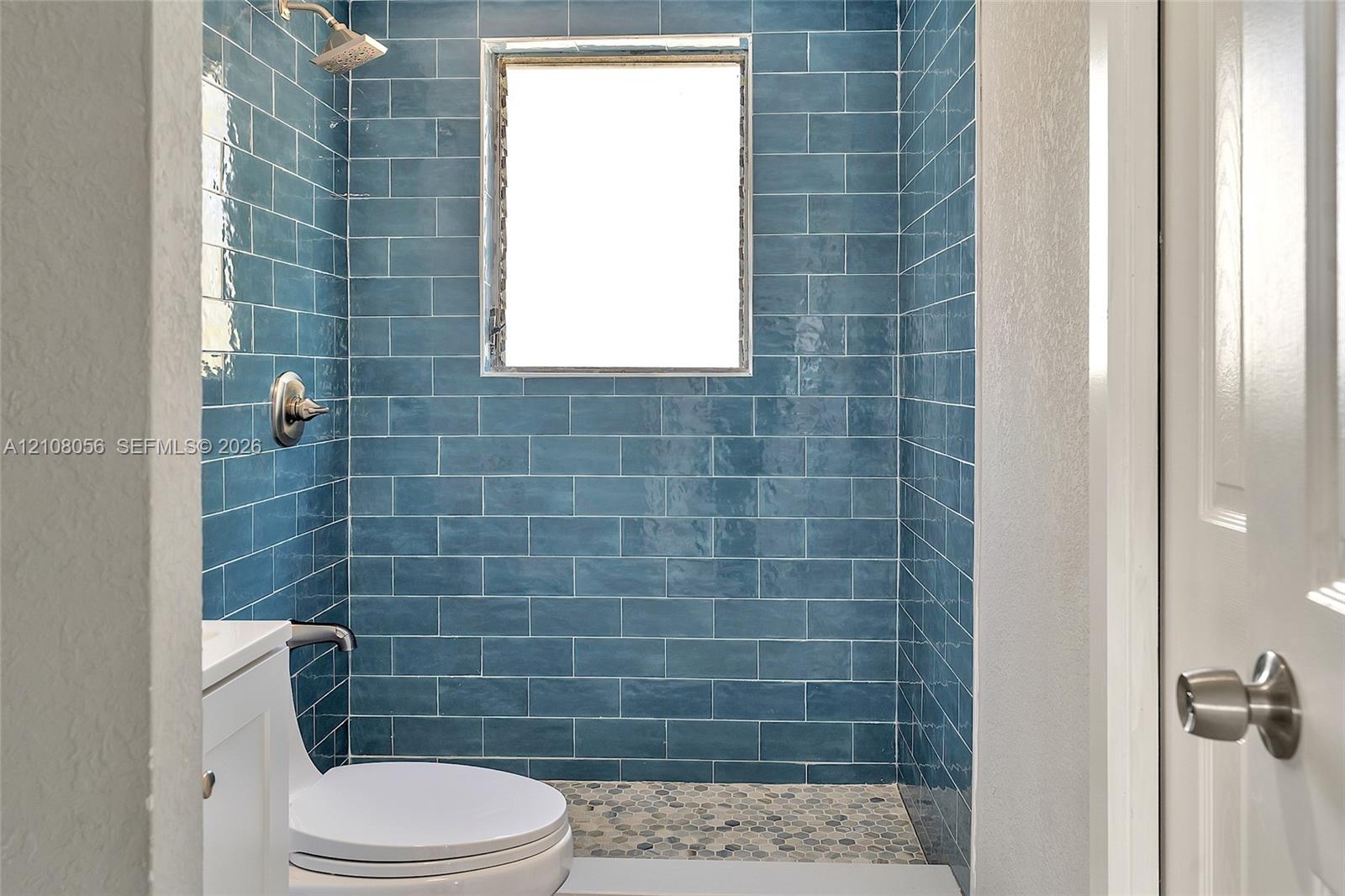 Walk-in shower featuring glossy blue subway tile, a natural stone pebble floor, a built-in shower head, and a privacy window