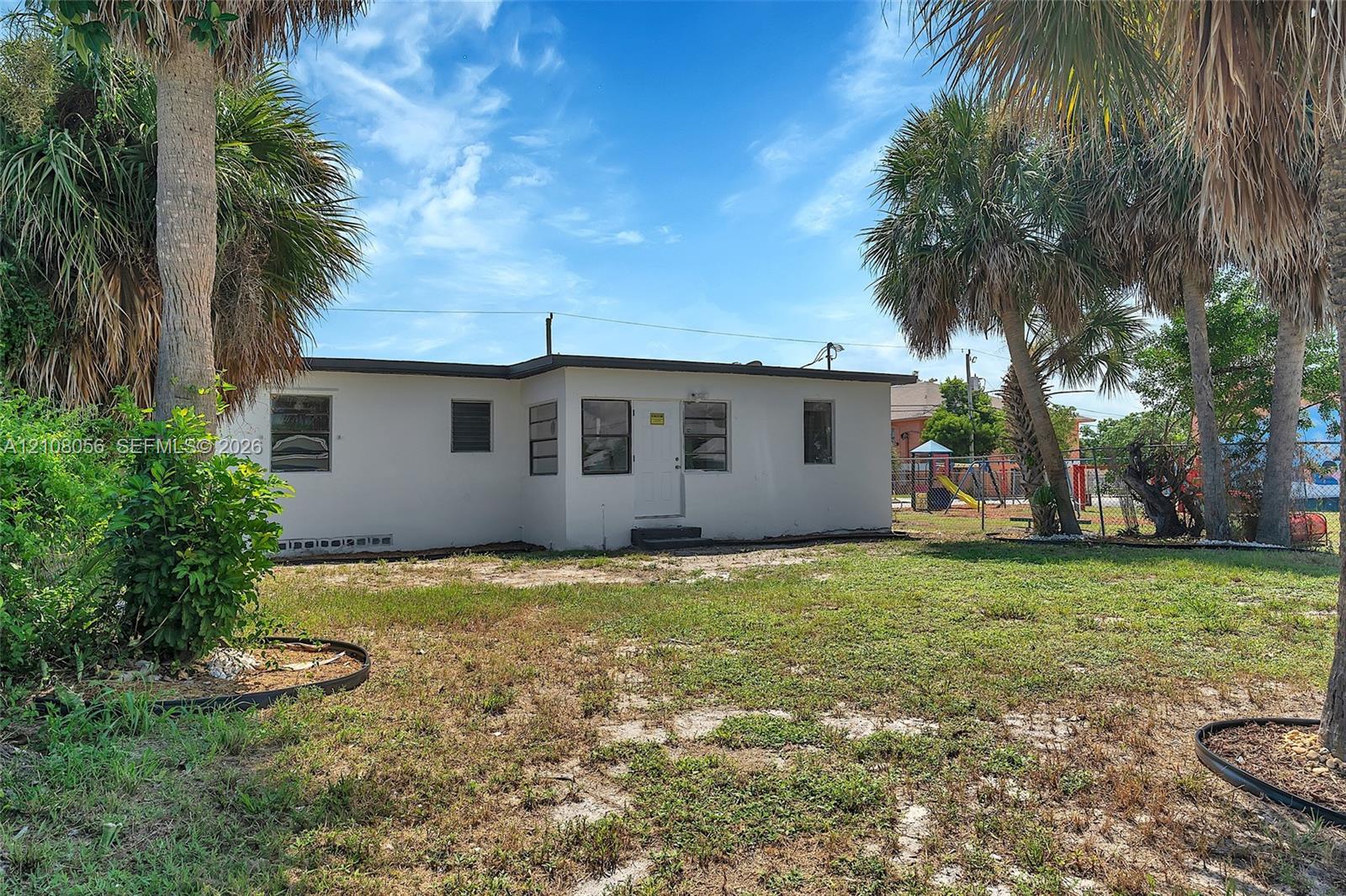 Exterior of a single-story dwelling featuring a white stucco facade, multi-pane windows, and a level yard with mature palm trees