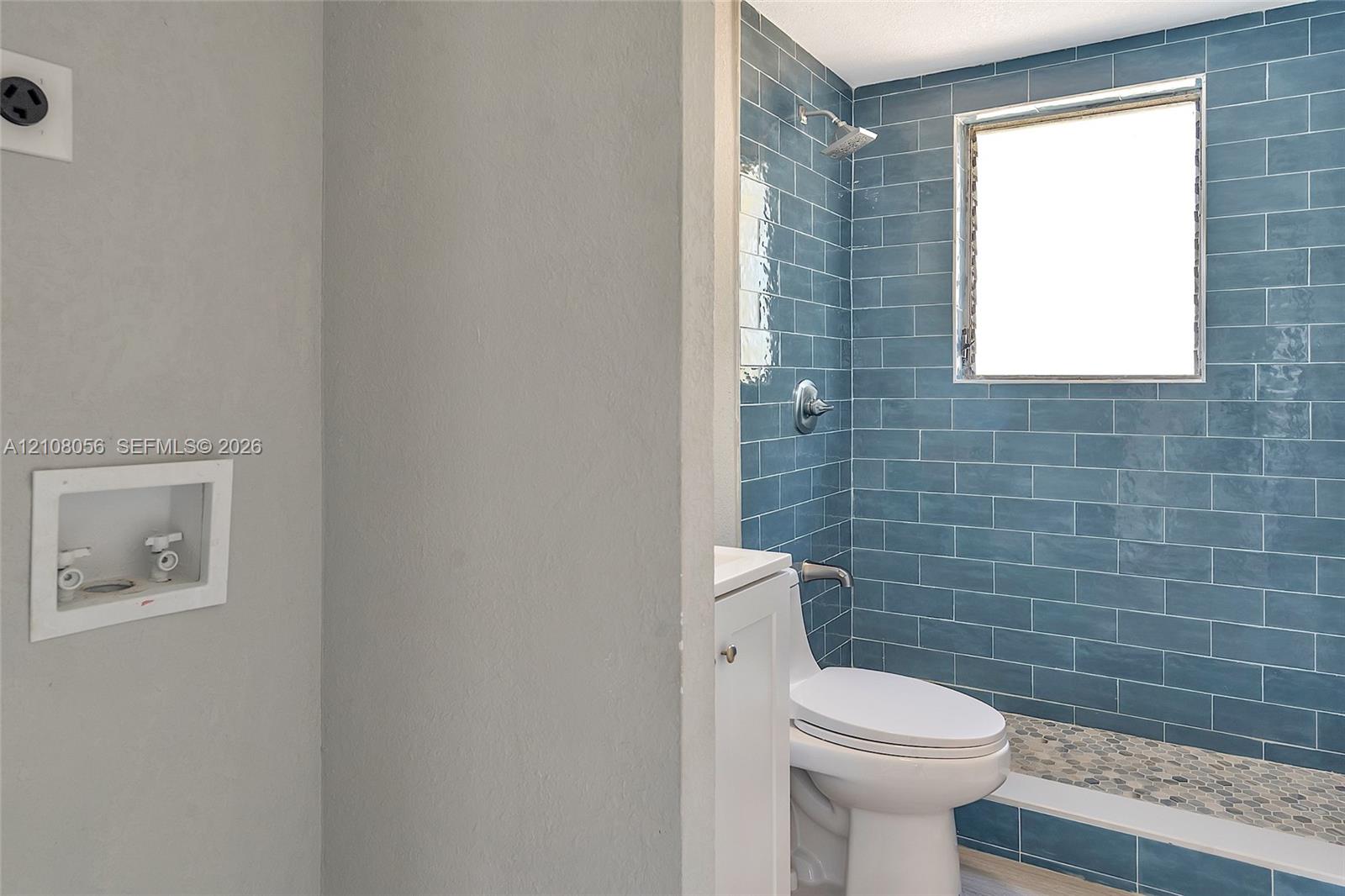 Bathroom featuring blue subway wall tile, pebble-stone shower flooring, a wall-mounted shower fixture, an integrated laundry hookup box, and a white pedestal vanity