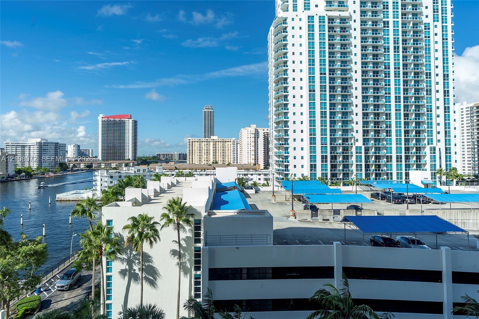 High-rise residential exterior with multi-level parking structure, blue canvas shade coverings, palm-lined perimeter, and waterfront access