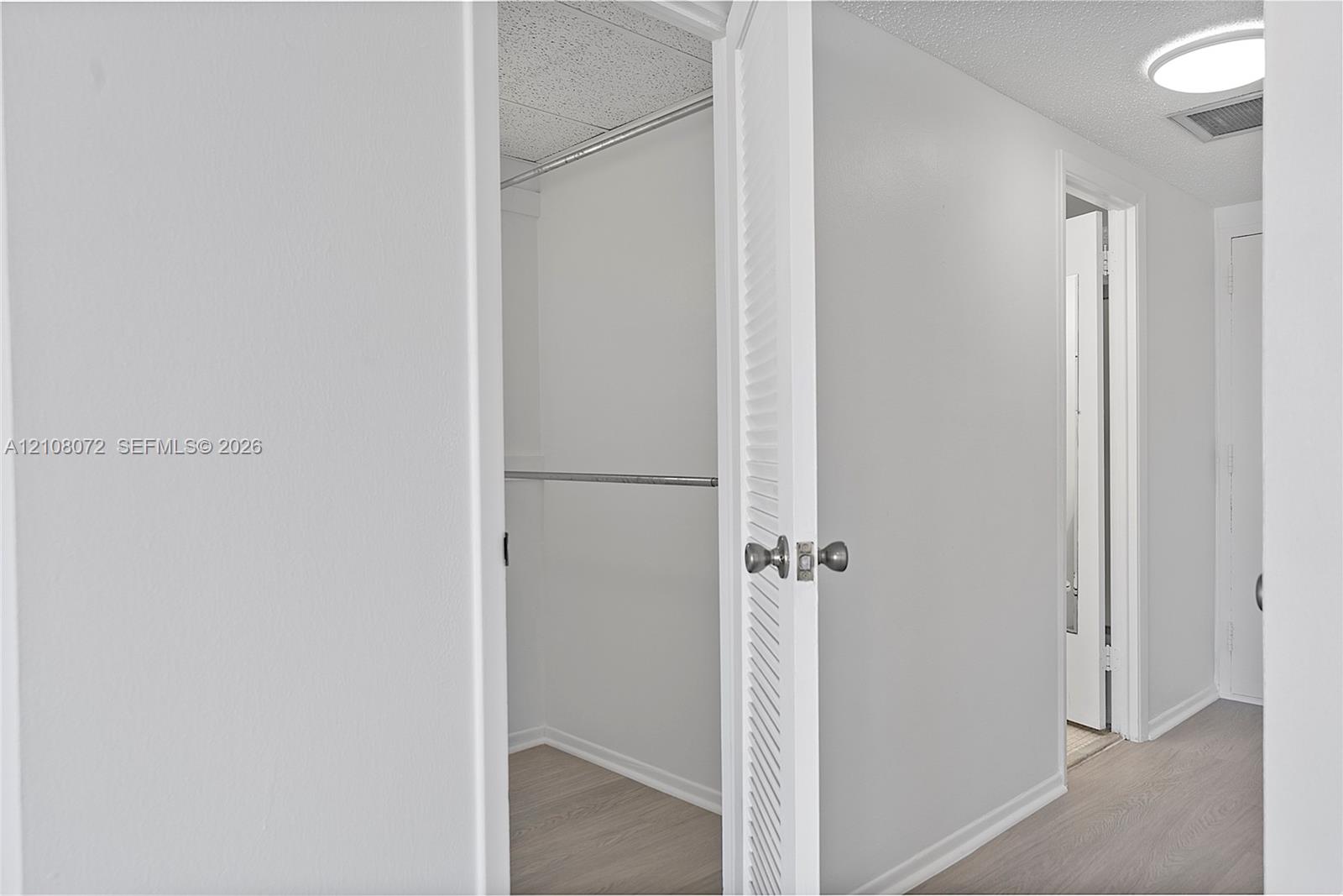 Walk-in closet featuring light-toned wood-finish flooring, white walls, and a metal hanging rod, complemented by a white louvered door with a brushed metallic knob