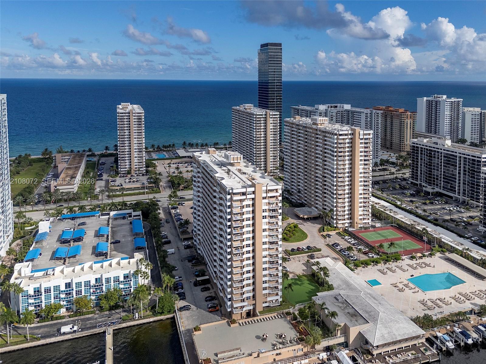 Aerial perspective of coastal residential complex featuring outdoor swimming pool, tennis court, boat slips, and parking structures