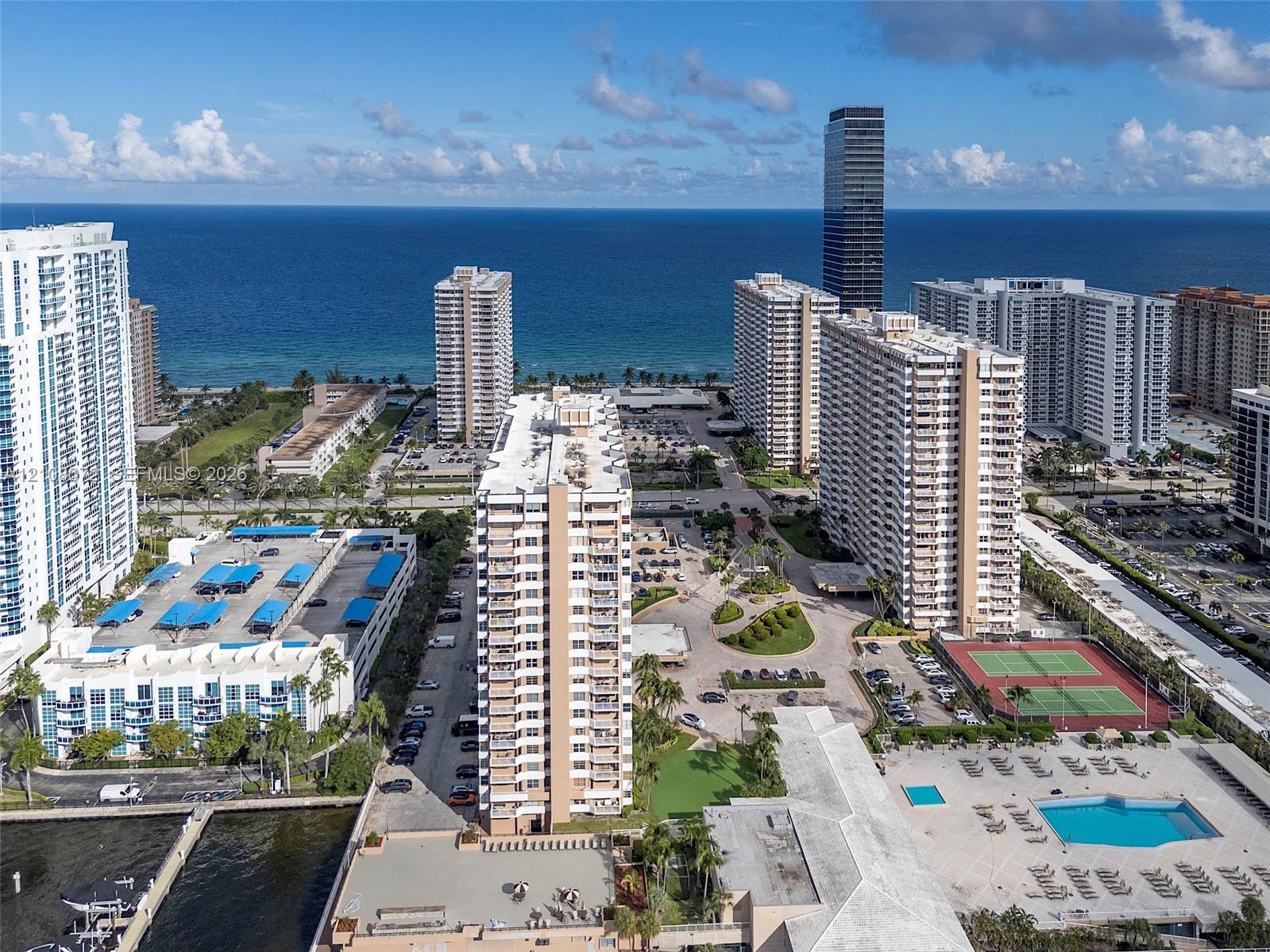 Aerial perspective featuring waterfront residential buildings, outdoor swimming pool, hard-surfaced tennis courts, and landscaped green space