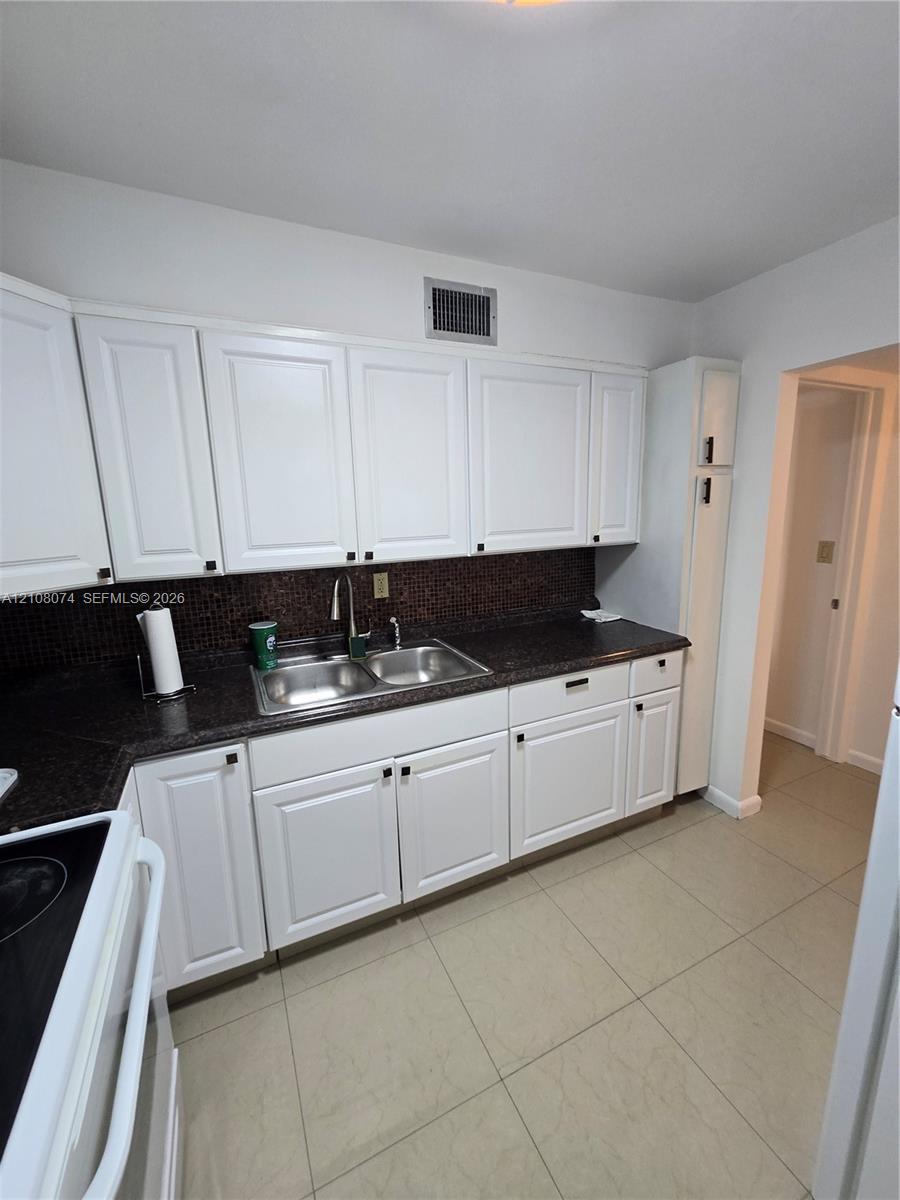 Kitchen featuring white cabinetry, dark countertops, a double-basin stainless steel sink, and light-toned tile flooring