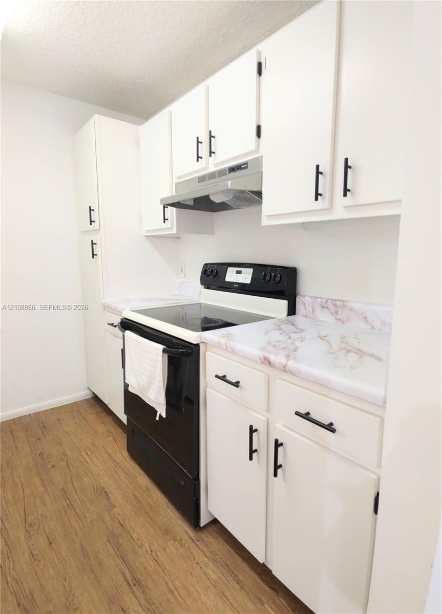 Kitchen featuring white cabinetry with matte black hardware, stone-patterned laminate countertops, stainless steel range hood, and wood-finish flooring