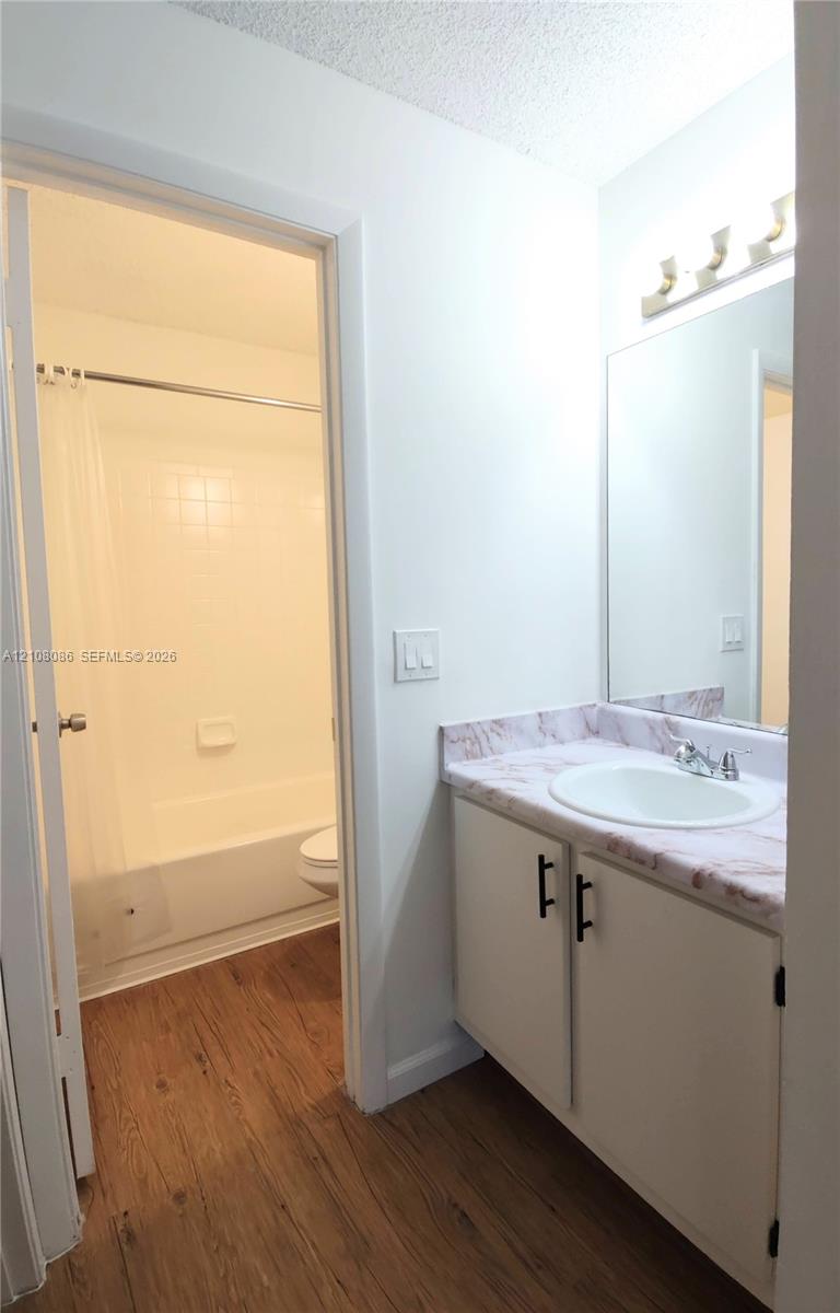 Bathroom featuring a vanity with white cabinetry, matte black hardware, and a stone-patterned countertop with an integrated basin, paired with wood-finish flooring and a wall-mounted light fixture above the mirror