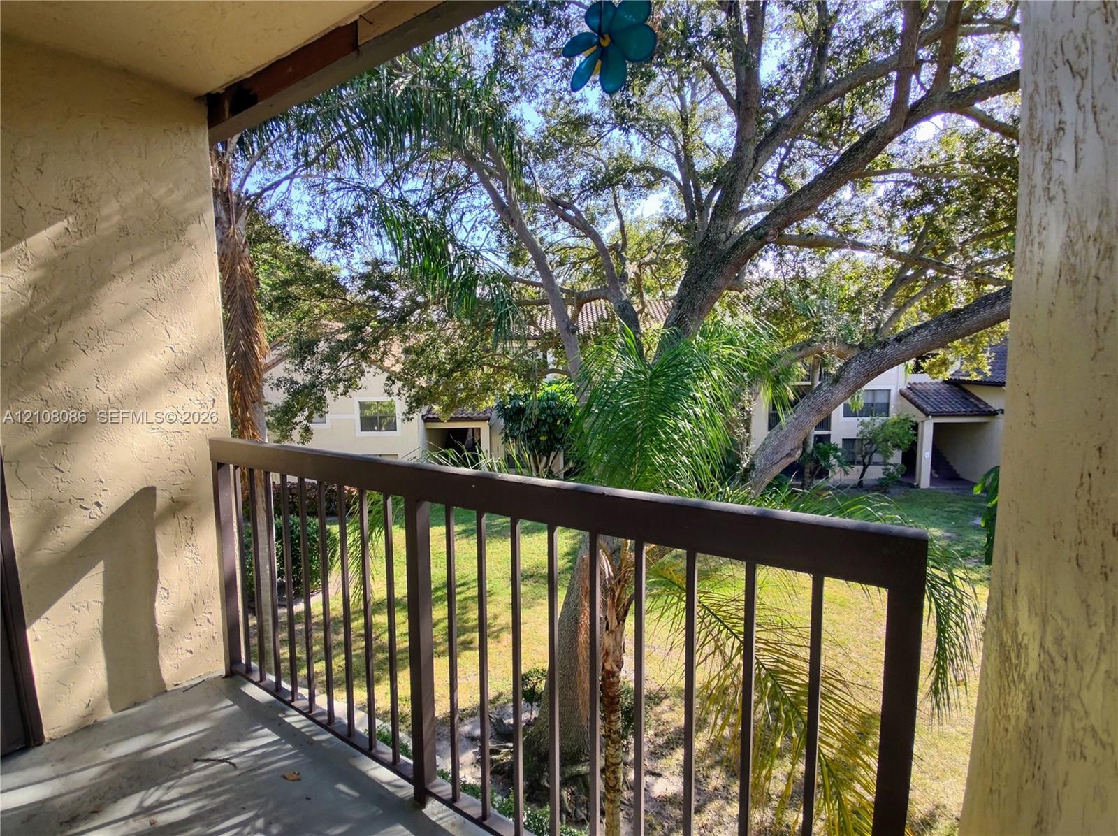 Covered balcony with metal railing, stucco walls, and mature landscaping