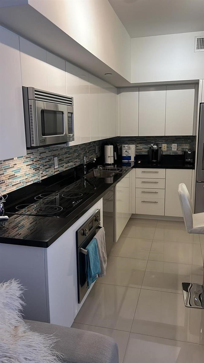 Kitchen featuring high-gloss white cabinetry, polished dark stone countertops, linear glass mosaic tile backsplash, integrated stainless steel appliances, and large-format tile flooring