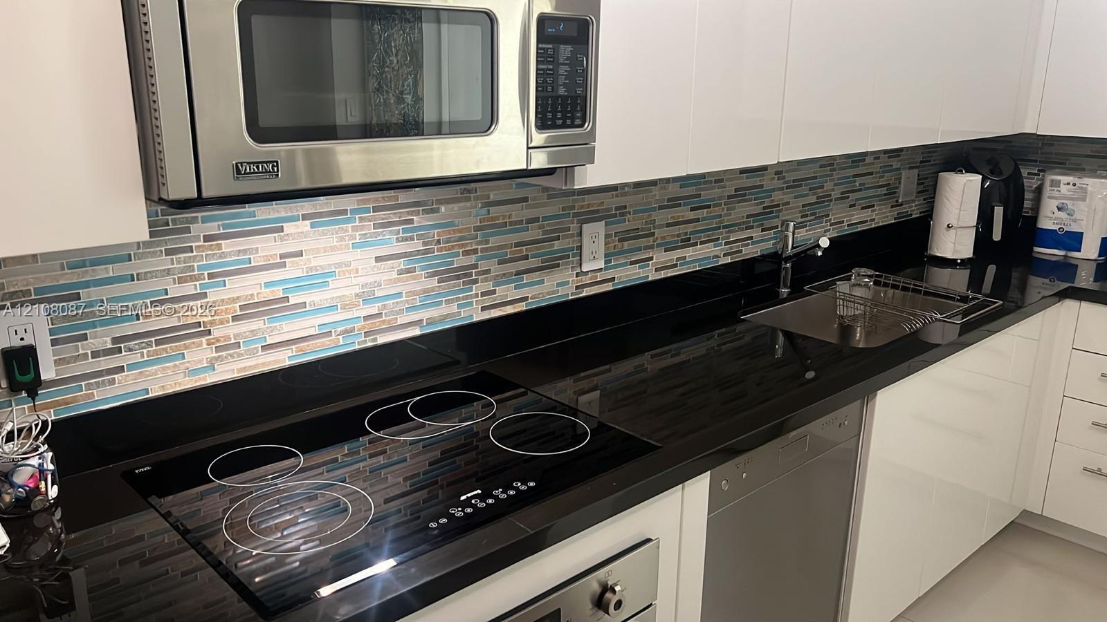 Kitchen featuring white flat-panel cabinetry, dark solid-surface countertops, stainless steel microwave, glass-top electric range, and mosaic tile backsplash
