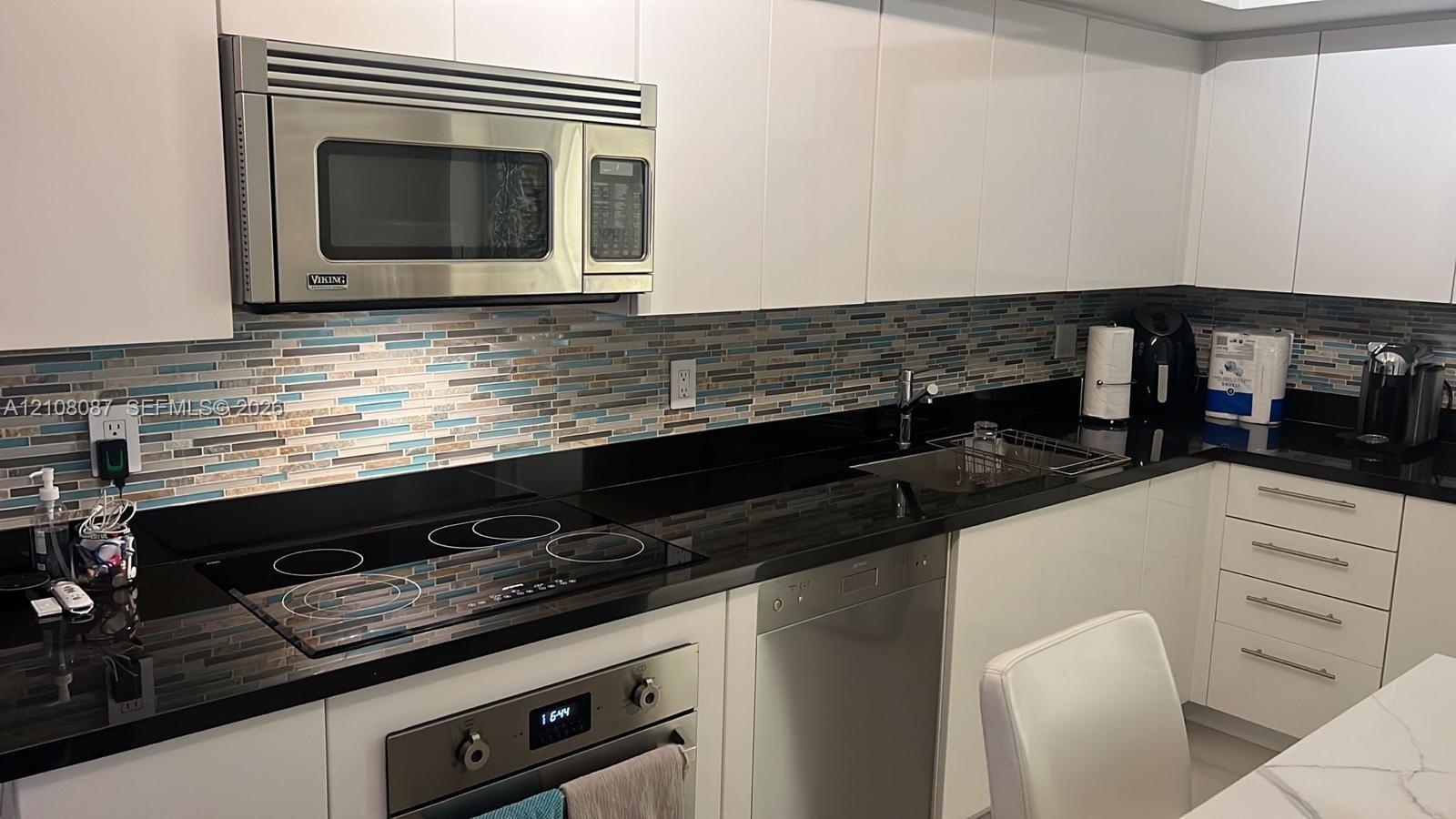 Kitchen featuring white slab cabinetry, black countertops, multi-toned glass tile backsplash, stainless steel built-in microwave, and electric cooktop