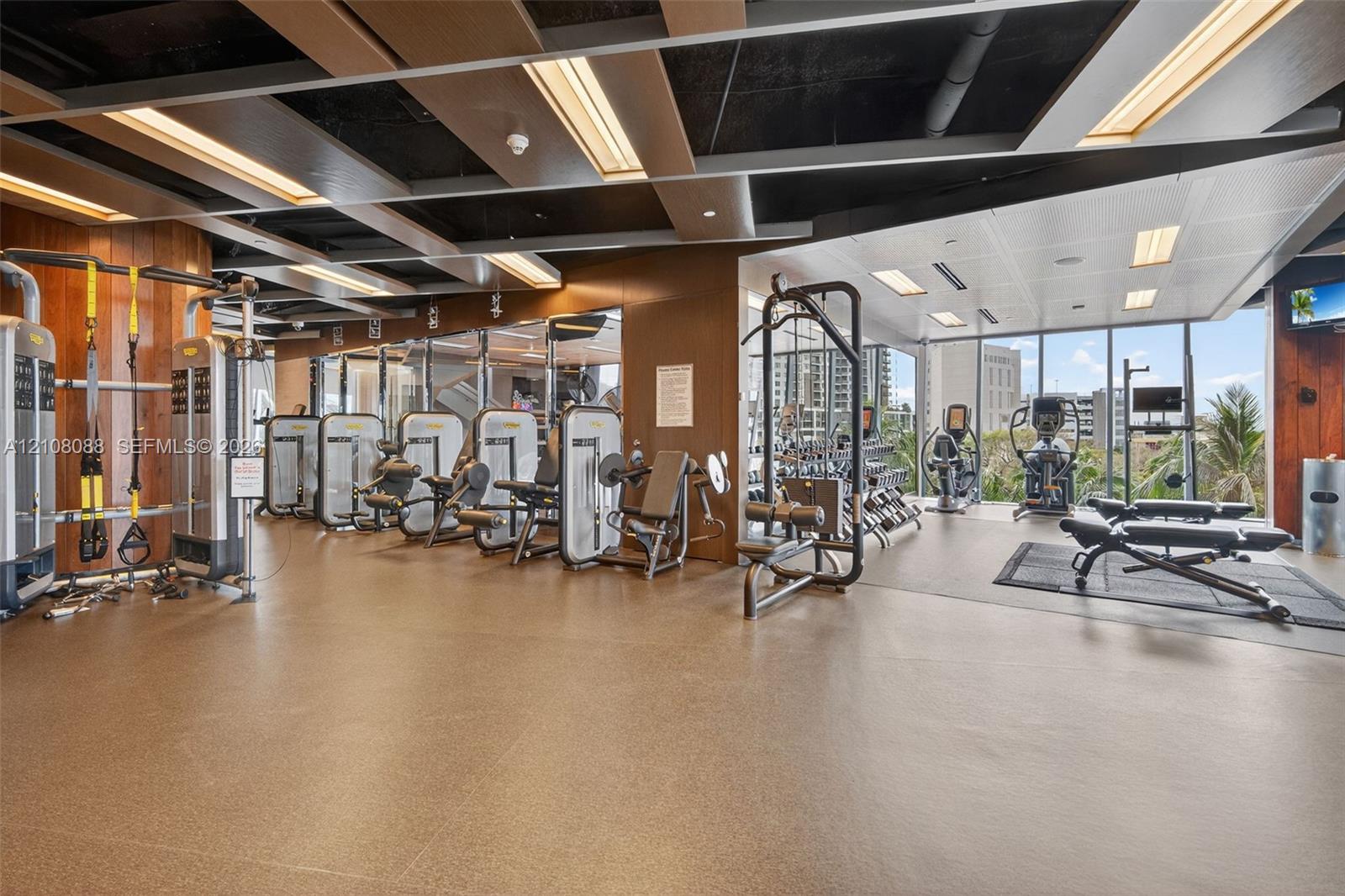 Fitness center featuring floor-to-ceiling windows, high-contrast coffered ceiling, recessed linear lighting, and wood-paneled accent walls