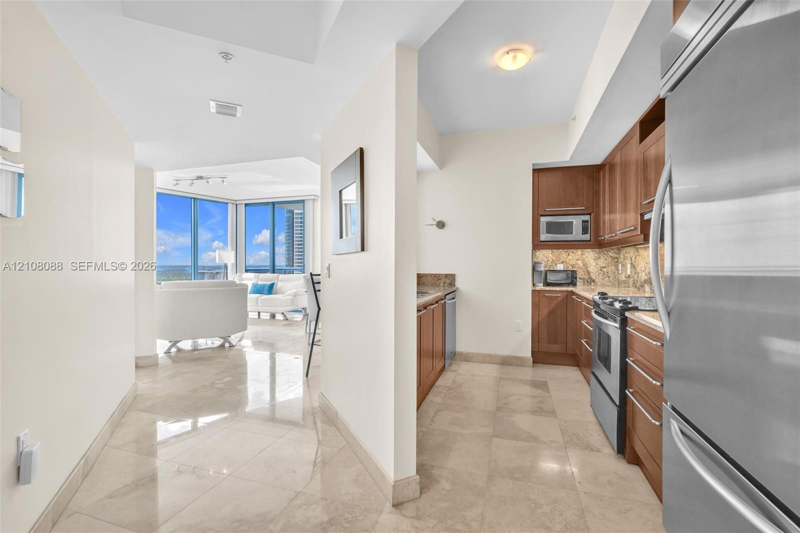 Entry and kitchen featuring wood cabinetry, stone countertops, stone flooring, and stainless steel appliances, with floor-to-ceiling windows Entry and kitchen featuring wood cabinetry, stone countertops, stone flooring, and stainless steel appliances, with floor-to-ceiling windows