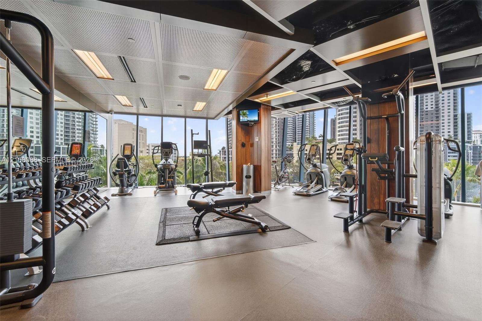 Fitness center featuring floor-to-ceiling windows, rubberized flooring, wood-paneled pillars, and a grid-style dropped ceiling with recessed lighting