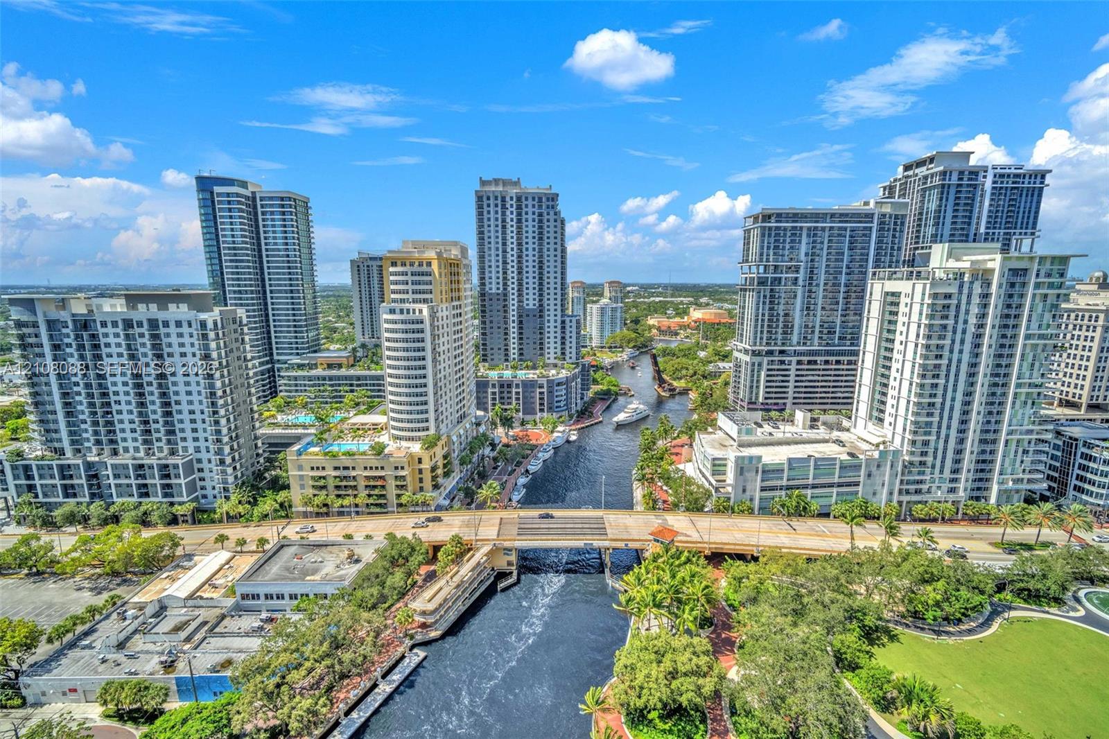 Aerial perspective of a high-density urban corridor featuring a central waterway, bridge infrastructure, mature landscaping, and numerous mid-rise and high-rise structures Aerial perspective of a high-density urban corridor featuring a central waterway, bridge infrastructure, mature landscaping, and numerous mid-rise and high-rise structures