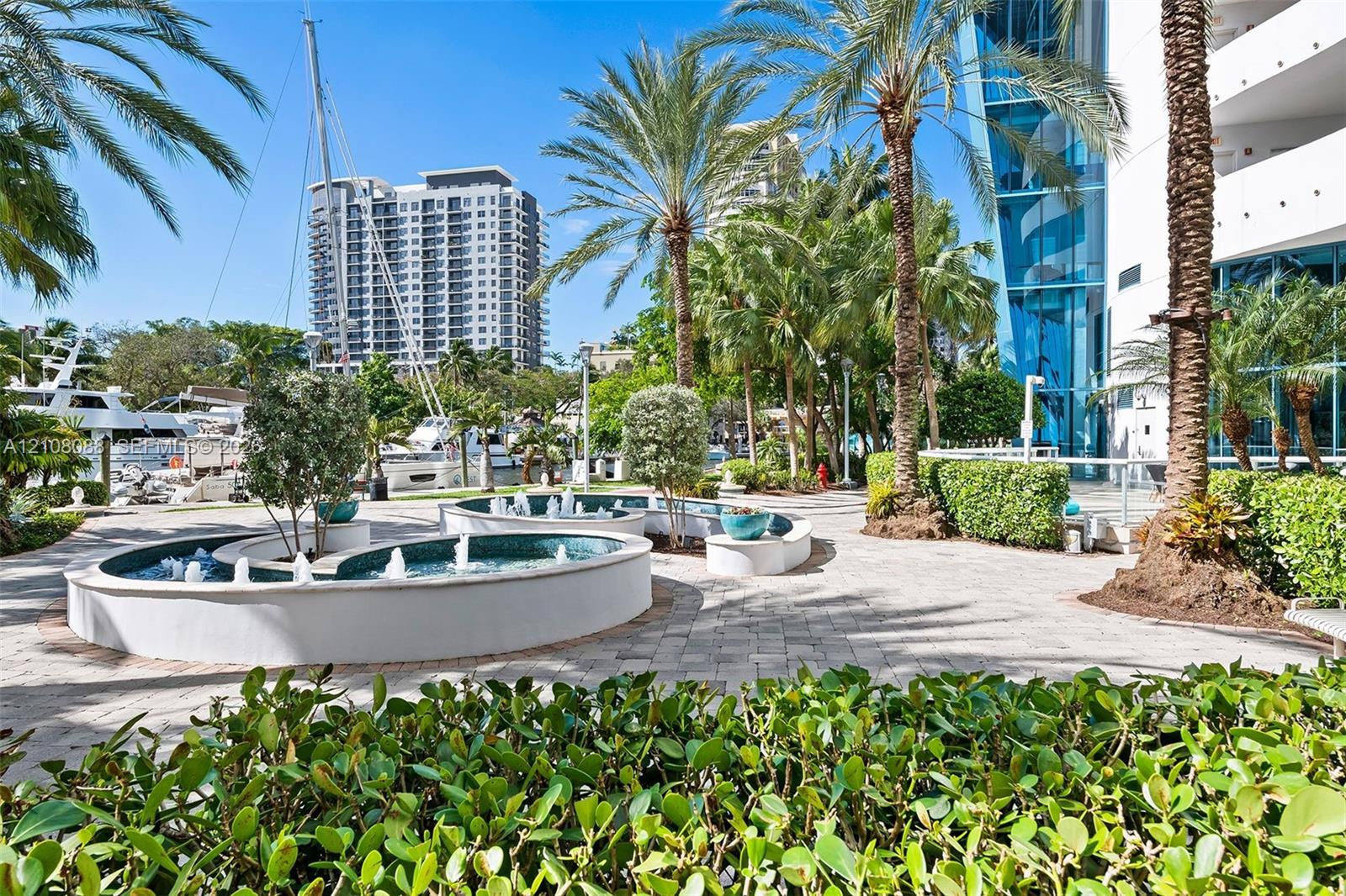 Outdoor courtyard featuring tiered water fountains, stone paver pathways, mature palm trees, and manicured hedges