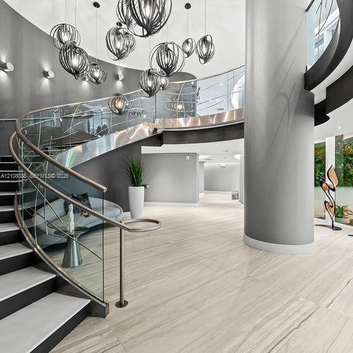 Curved staircase featuring glass railings with metal handrails, large-format tile flooring, a multi-pendant light installation, and a floor-to-ceiling structural column