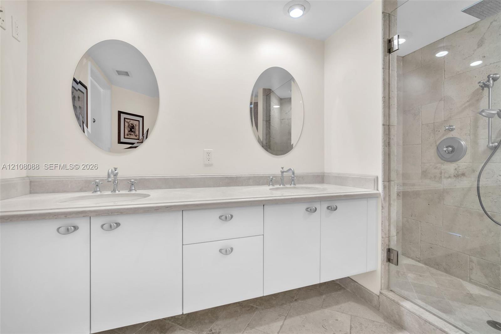 Dual-sink vanity with white cabinetry, light-toned stone-finish countertop, dual oval mirrors, and a glass-enclosed shower featuring stone wall tiles