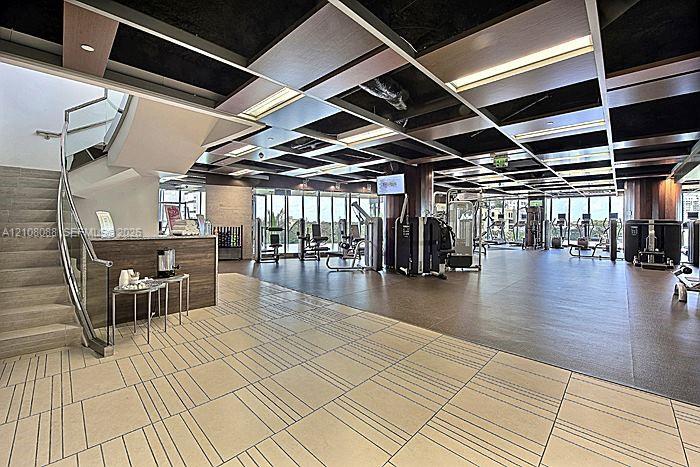 Fitness center featuring a coffered ceiling with recessed lighting, full-height glass walls, a wood-finish staircase with a metal railing, and neutral-toned geometric floor tiling