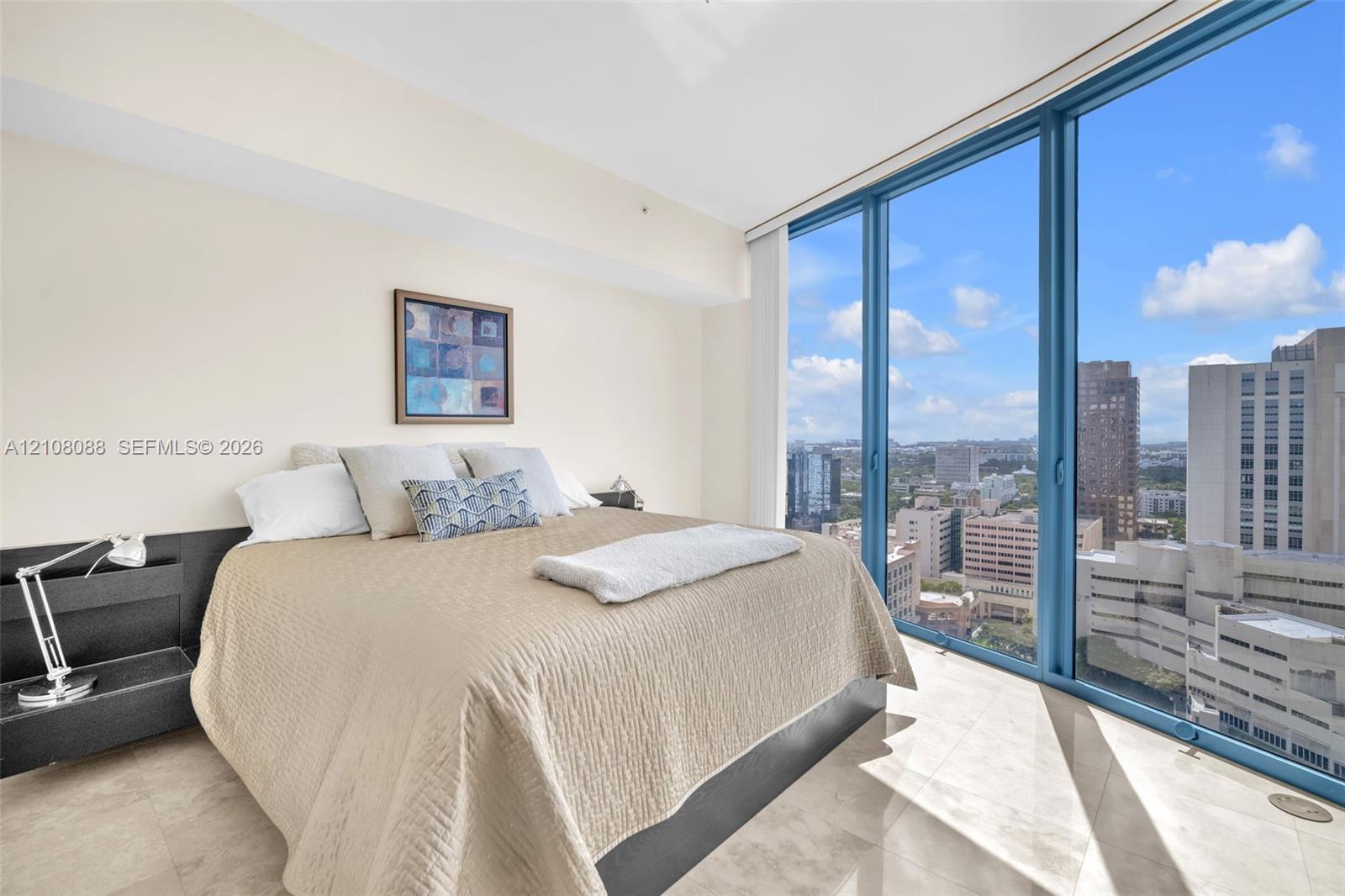 Floor-to-ceiling windows featuring urban scenery, expansive light-toned tile flooring, and a sleek, built-in dark finish headboard