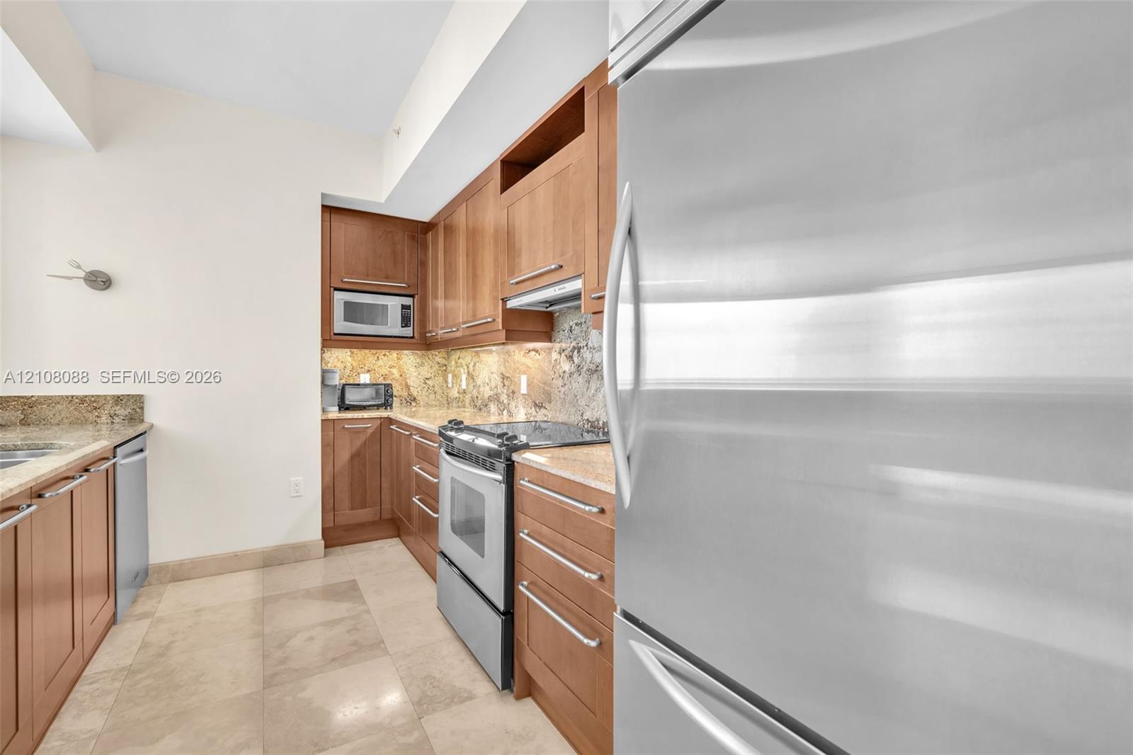 Kitchen featuring natural wood cabinetry, stone countertops, stone tile backsplash, stainless steel appliances, and large-format tile flooring Kitchen featuring natural wood cabinetry, stone countertops, stone tile backsplash, stainless steel appliances, and large-format tile flooring