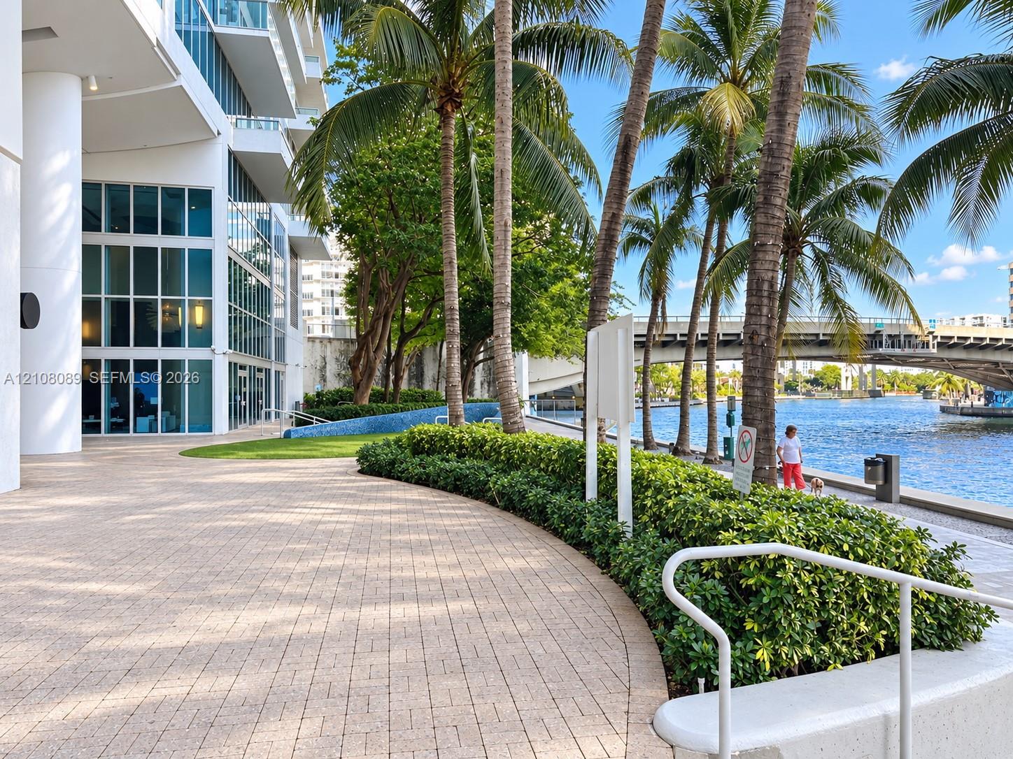 Waterfront walkway featuring paver hardscaping, mature palm trees, manicured hedges, and floor-to-ceiling glass building facade