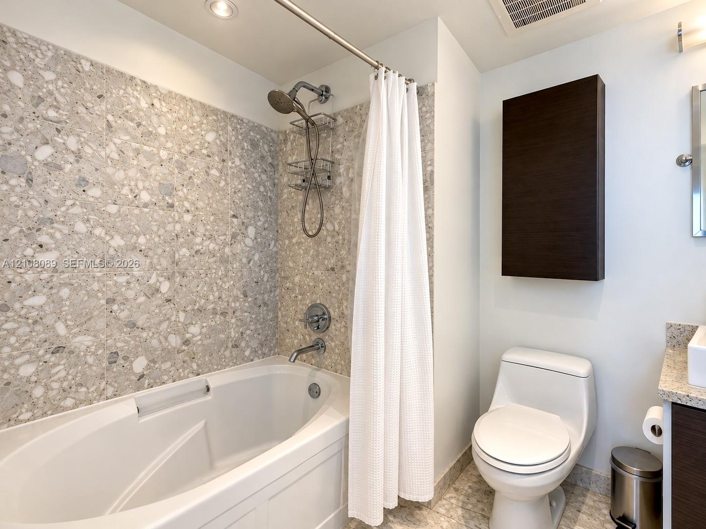 Bathroom featuring a soaking tub with stone-tiled surround, handheld shower fixture, dual-flush toilet, wall-mounted storage cabinet, and vanity with stone countertop