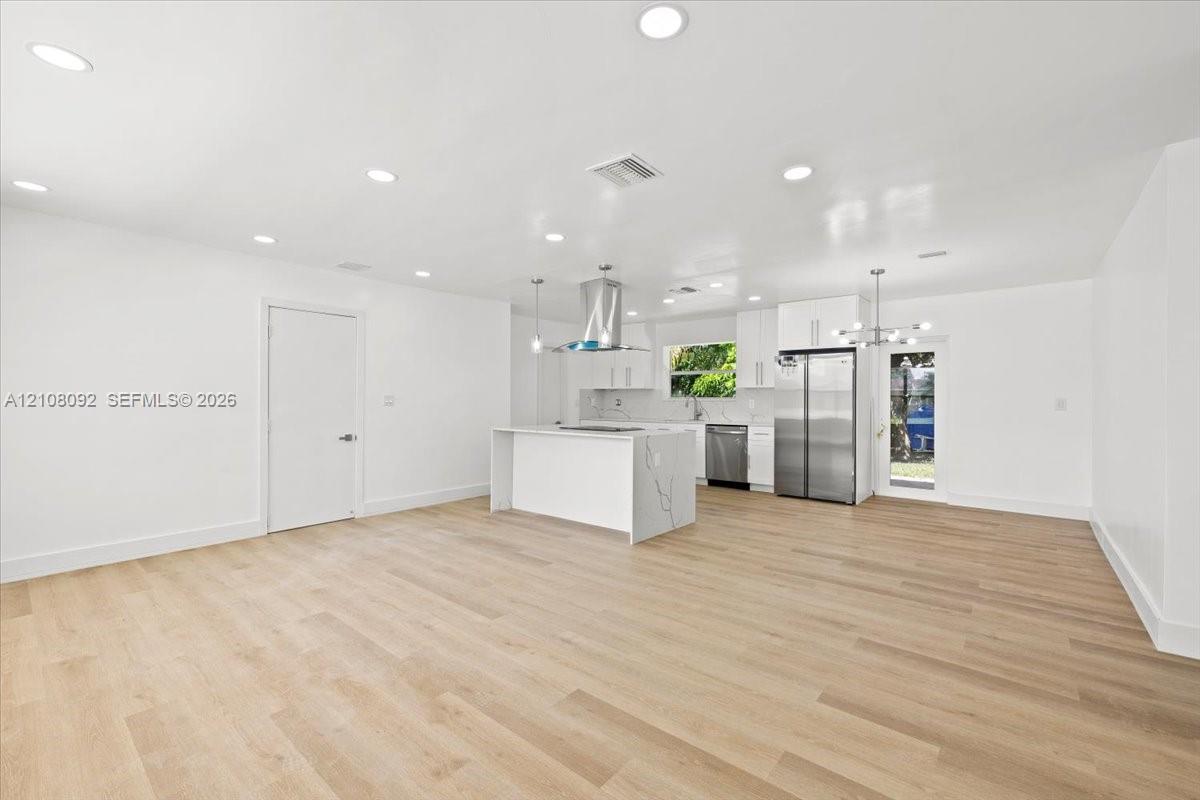 Open-concept interior featuring light wood-finish flooring, recessed lighting, a central island with stone-finish waterfall countertop, stainless steel appliances, and white shaker-style cabinetry