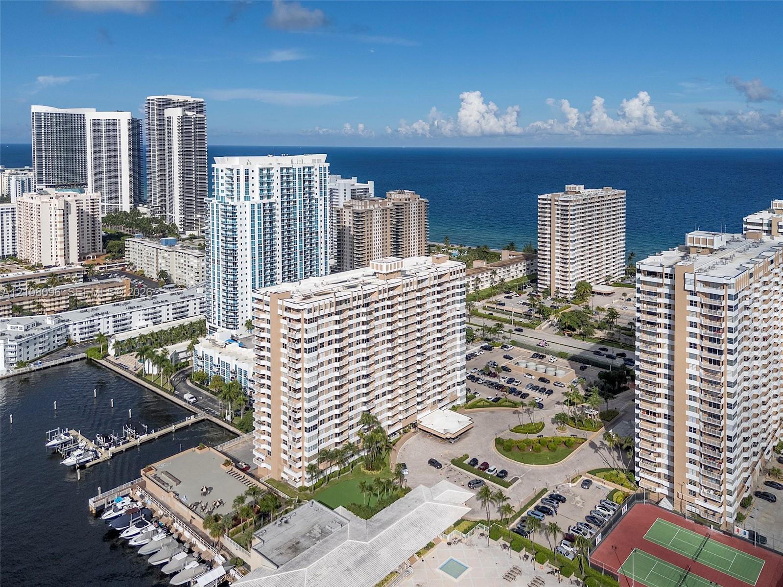 Aerial perspective of a waterfront high-rise complex featuring deep-water boat slips, paved parking facilities, multi-court tennis area, and proximity to oceanfront coastline