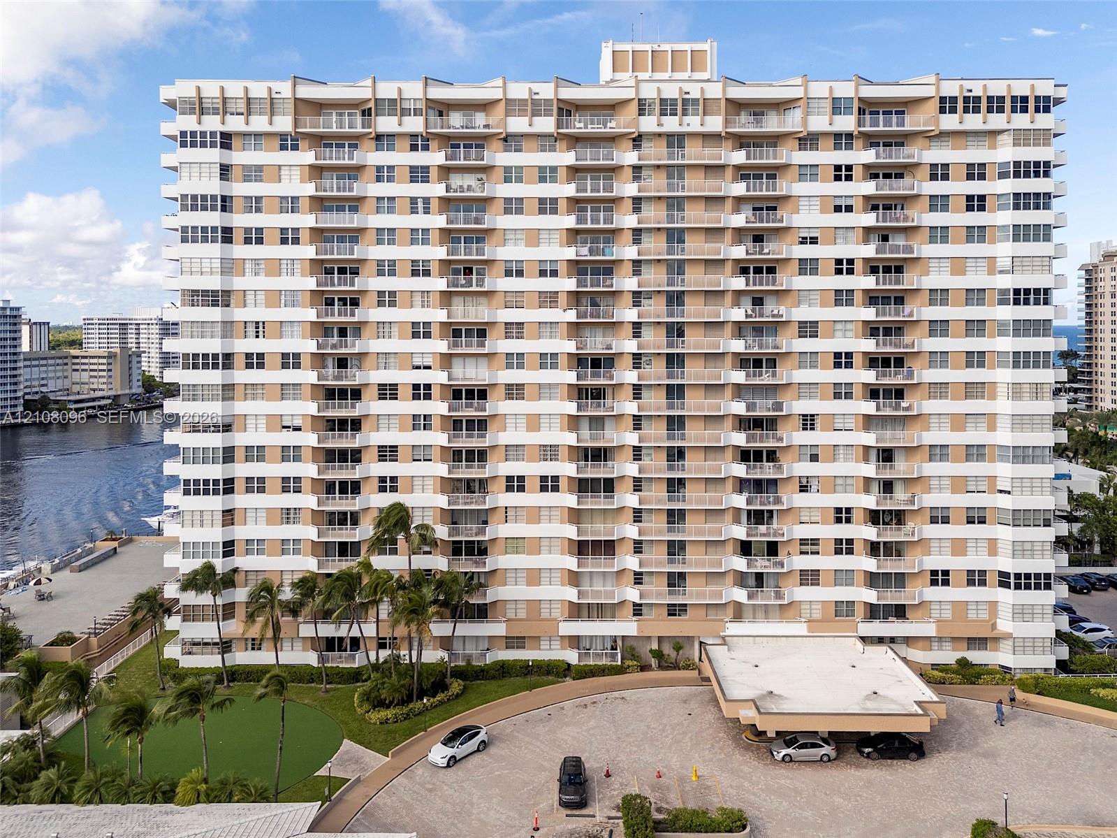High-rise residential building featuring stacked private balconies, a covered porte-cochre, paved motor court, and landscaped grounds with mature palm trees