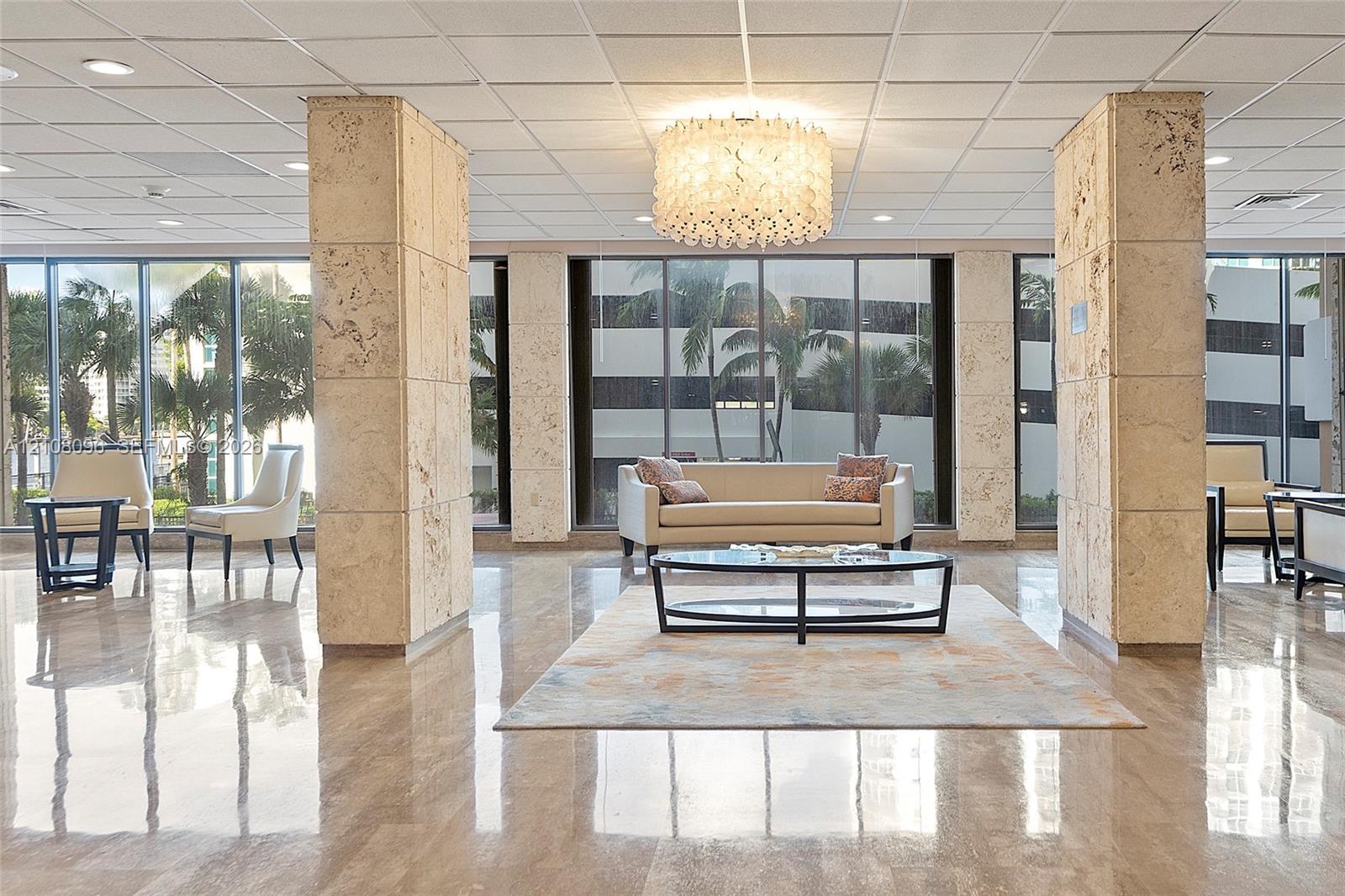 Spacious lobby featuring polished stone flooring, floor-to-ceiling windows, architectural stone-clad support pillars, a multi-tiered circular chandelier, and a drop ceiling