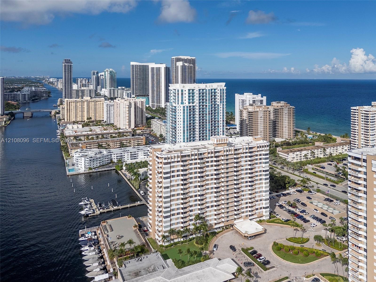 Aerial perspective of a multi-story residential building featuring integrated boat docking facilities, waterfront positioning, proximity to the coastline, and expansive paved parking areas