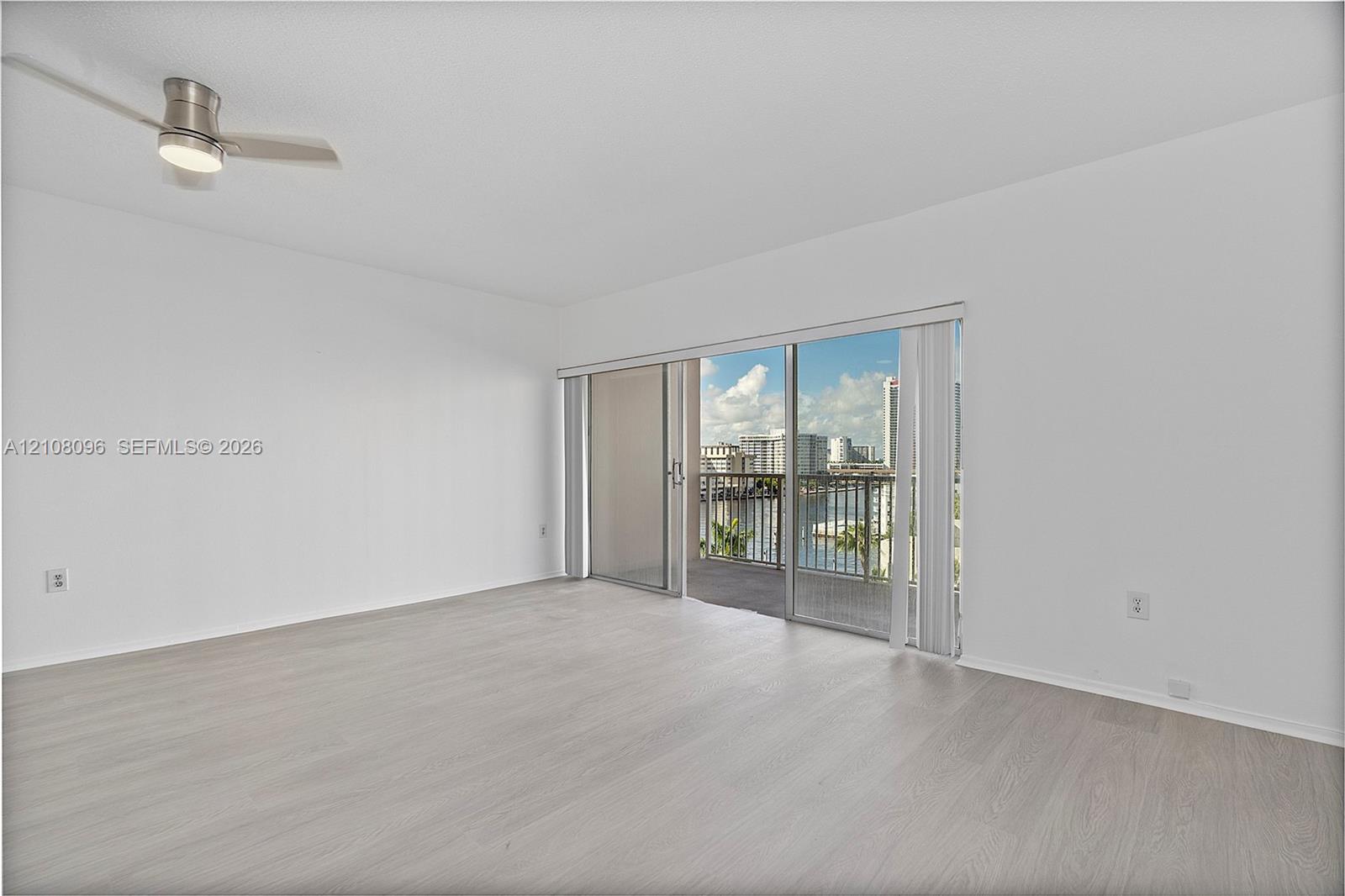 Neutral interior space featuring wood-finish flooring, a ceiling-mounted light fixture with fan, and a large sliding glass door providing access to an exterior balcony