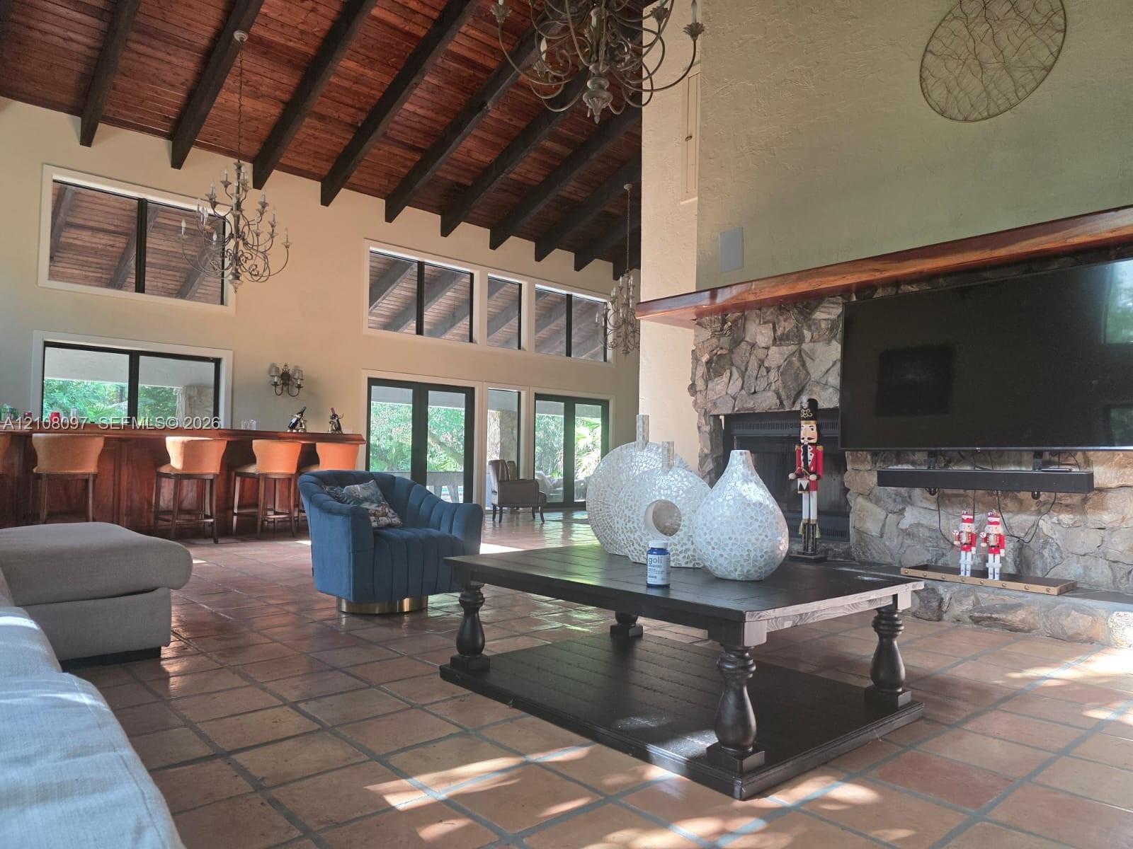 Vaulted ceiling with exposed wood beams, floor-to-ceiling stone fireplace, built-in wooden bar, and tile flooring