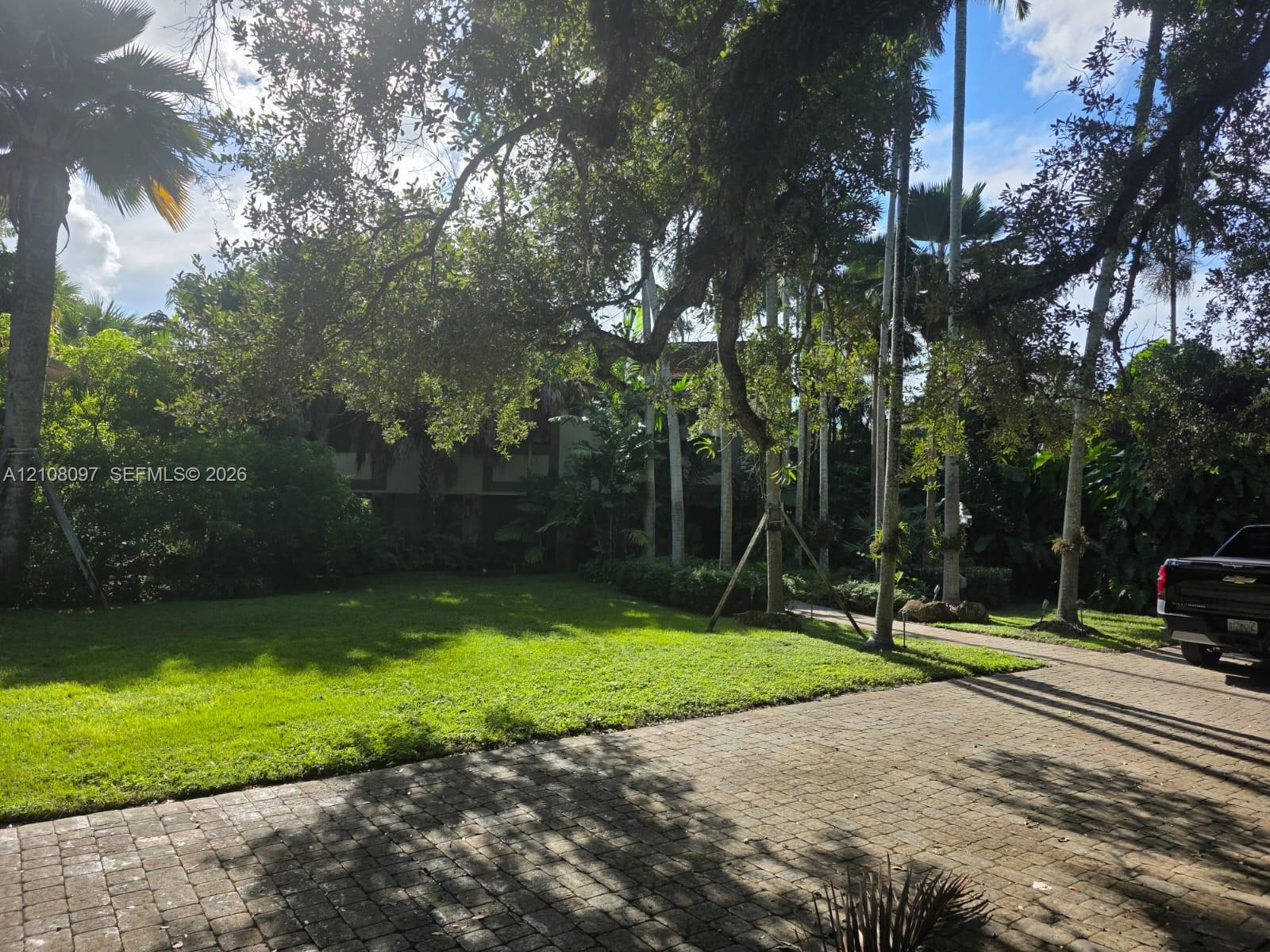 Expansive grounds featuring a paved brick driveway, mature canopy trees, manicured lawn, and established tropical landscaping