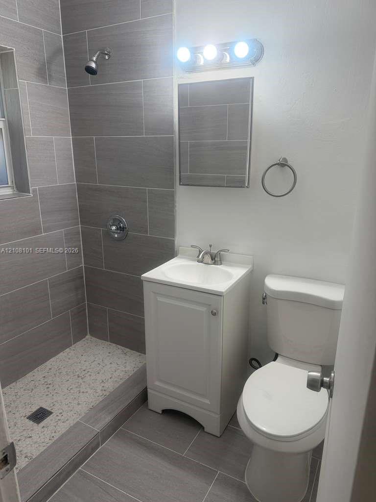 Bathroom featuring walk-in shower with large-format tile surround, integrated pebble-textured floor, white vanity cabinet with a ceramic sink, and vanity light fixture