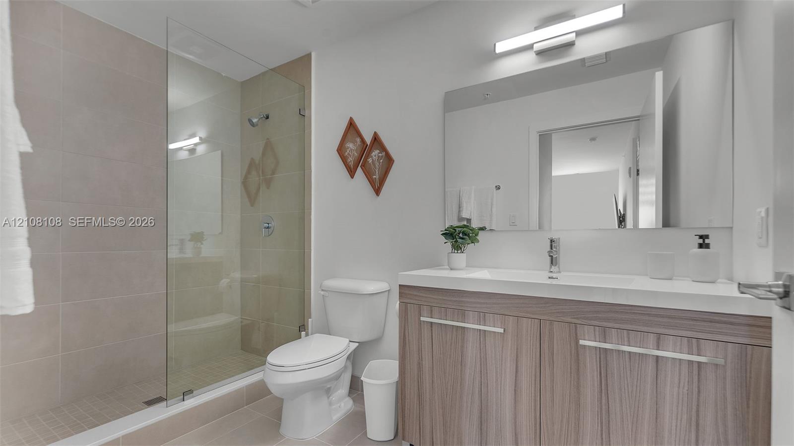 Bathroom featuring a wood-finish vanity with a white countertop, a frameless glass shower enclosure, oversized wall-mounted mirror, and linear light fixture Bathroom featuring a wood-finish vanity with a white countertop, a frameless glass shower enclosure, oversized wall-mounted mirror, and linear light fixture