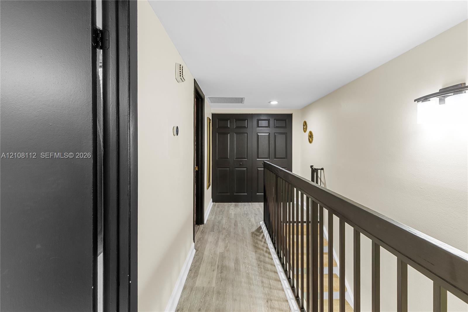 Hallway featuring wood-finish flooring, dark-toned interior doors, and a matching handrail with vertical balusters