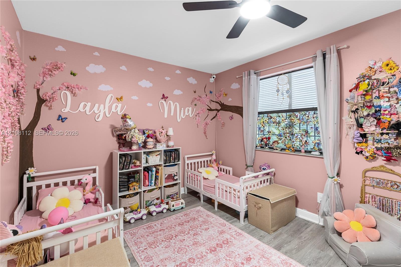 Wood-finish flooring, large window with horizontal blinds, and a multi-blade ceiling fan with integrated lighting