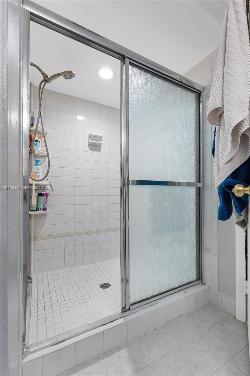 Shower enclosure featuring a chrome-framed sliding glass door, white tiled walls, a handheld shower fixture, and a built-in corner shelving unit