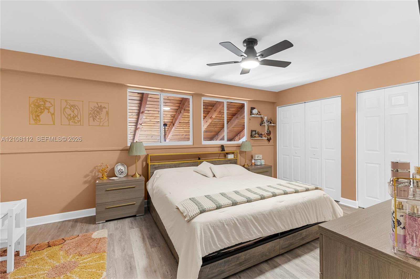 Bedroom featuring wood-finish flooring, dual windows with exposed wood rafters, ceiling fan with integrated lighting, and multiple bi-fold closets