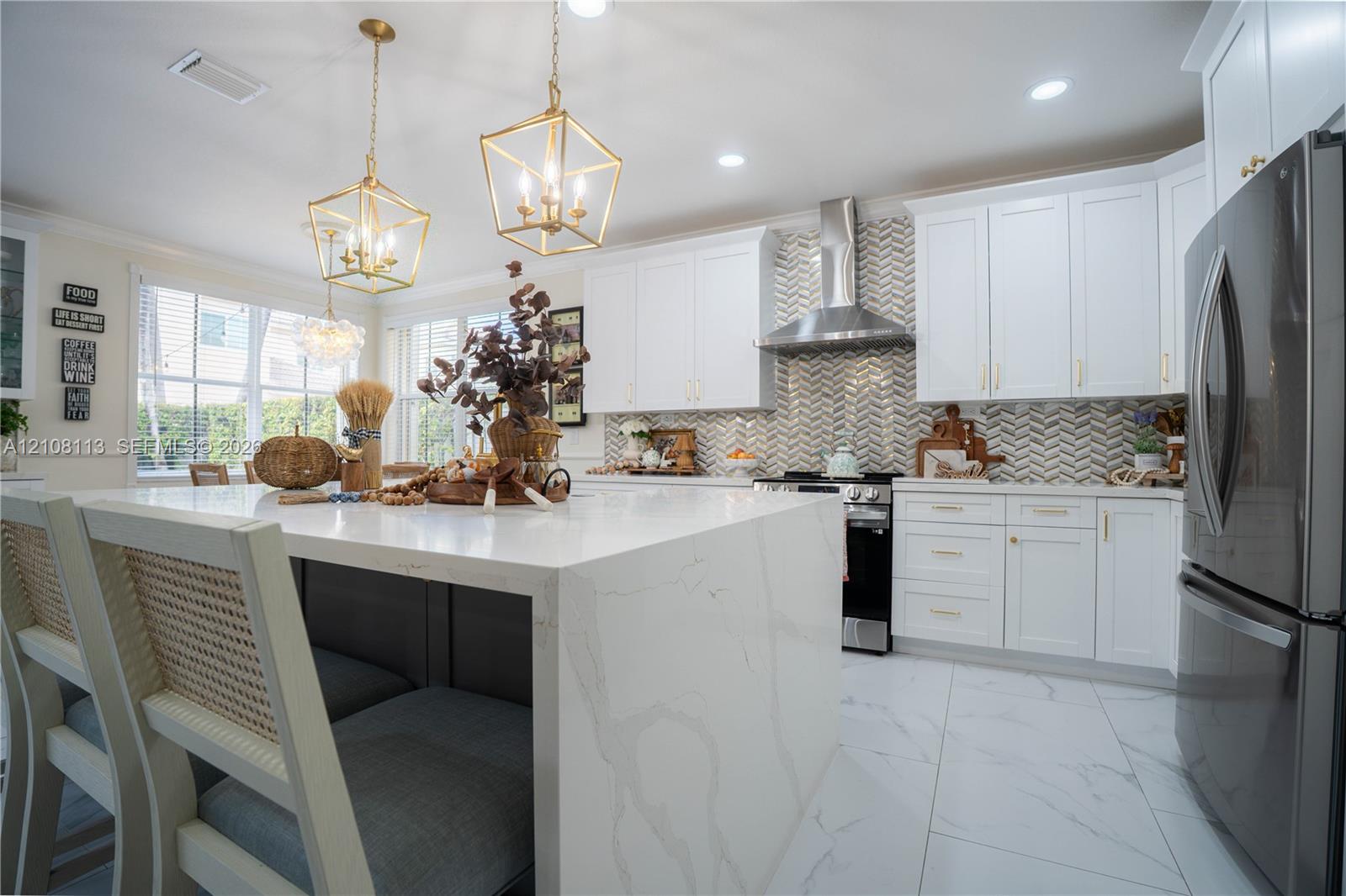 Kitchen featuring a waterfall-edge stone island, white shaker-style cabinetry, herringbone-patterned tile backsplash, stainless steel chimney-style range hood, and large-format tile flooring