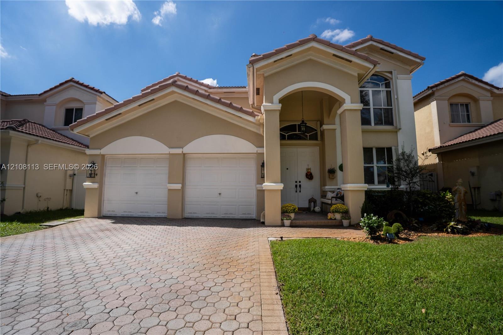 Two-story exterior featuring a stucco facade, tile roof, arched entryway with decorative columns, and a paved stone driveway leading to an attached two-car garage