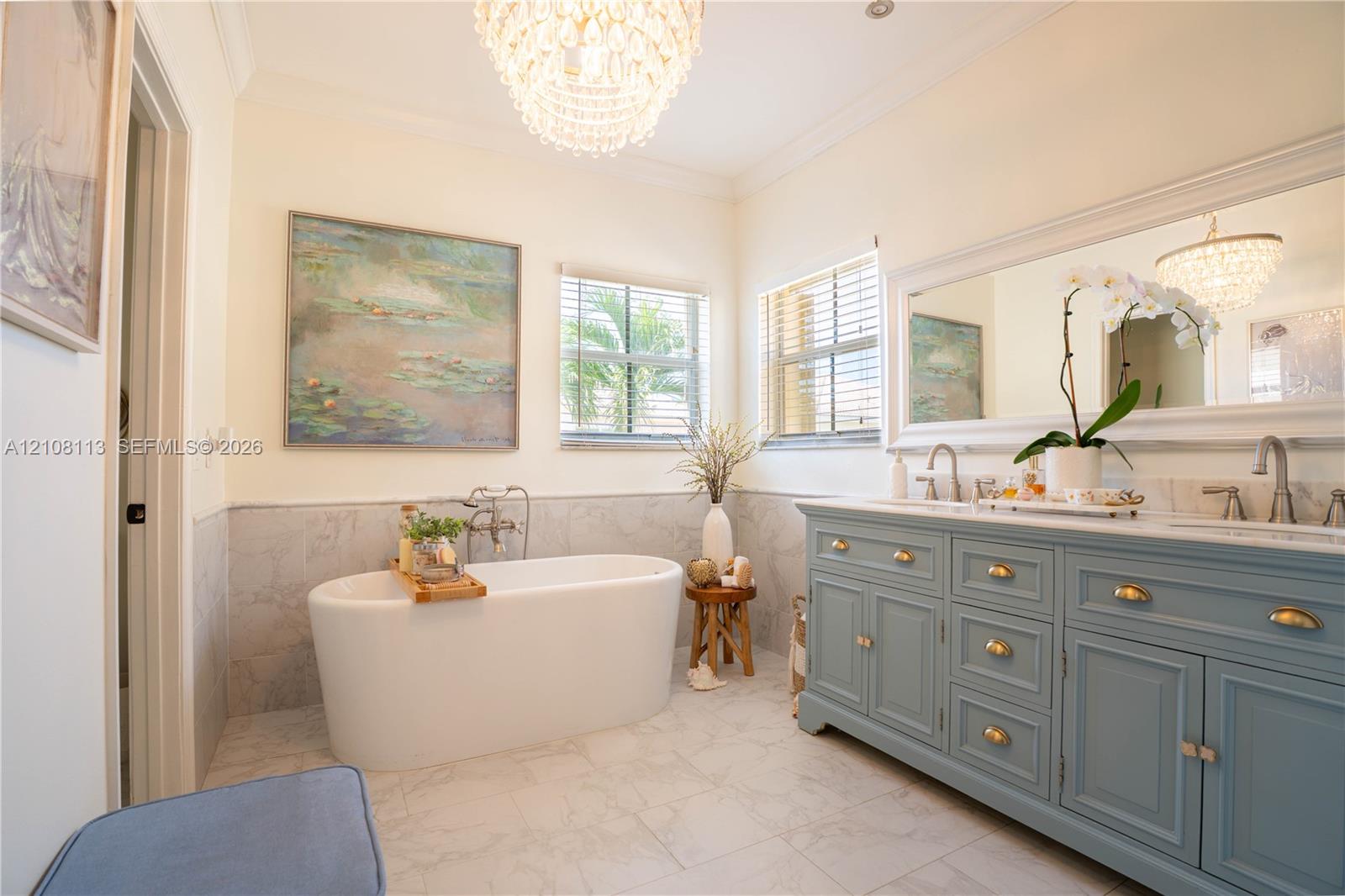 Freestanding soaking tub with wall-mounted plumbing, dual-sink vanity with brass-finish hardware, stone-finish flooring, half-wall tile wainscoting, and tiered crystal chandelier lighting