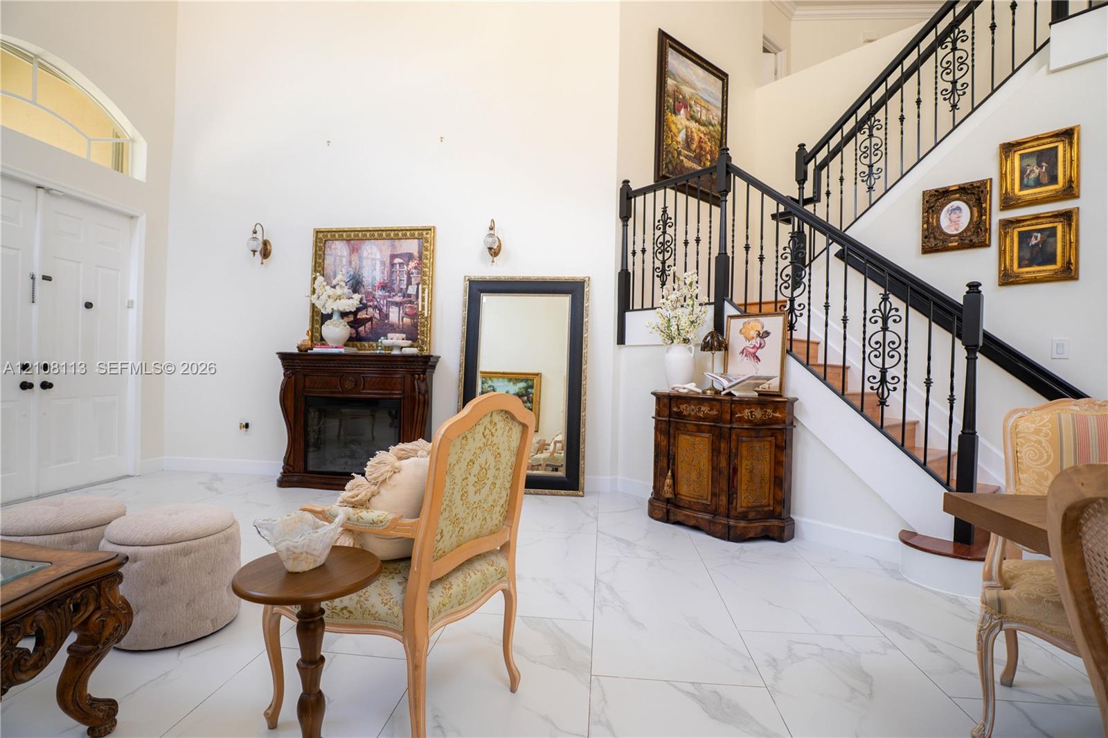 Grand entry featuring double front doors, vaulted ceilings, light-toned stone-finish tile flooring, a staircase with decorative wrought-iron railings, and an integrated dark wood-finish fireplace