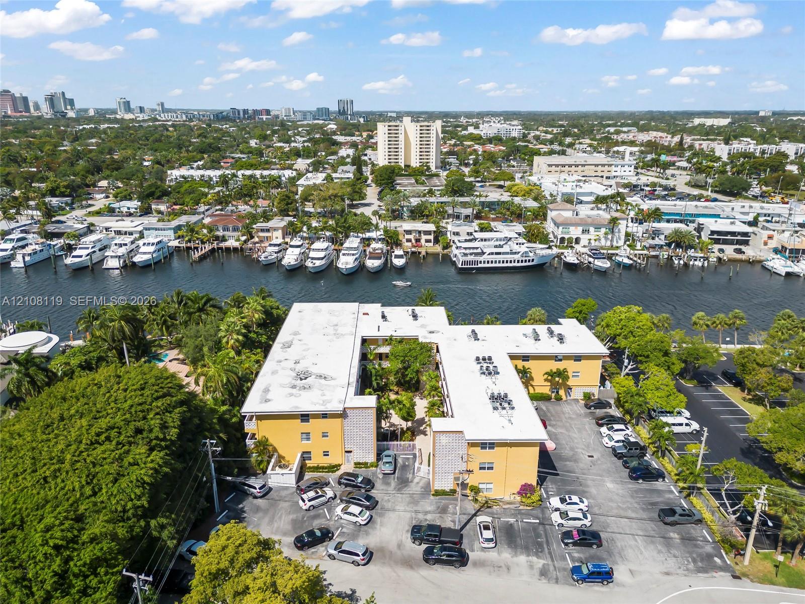 Aerial perspective of a multi-story building featuring a paved surface parking lot, exterior stucco finish, flat roofing, and proximity to a waterway with boat slips
