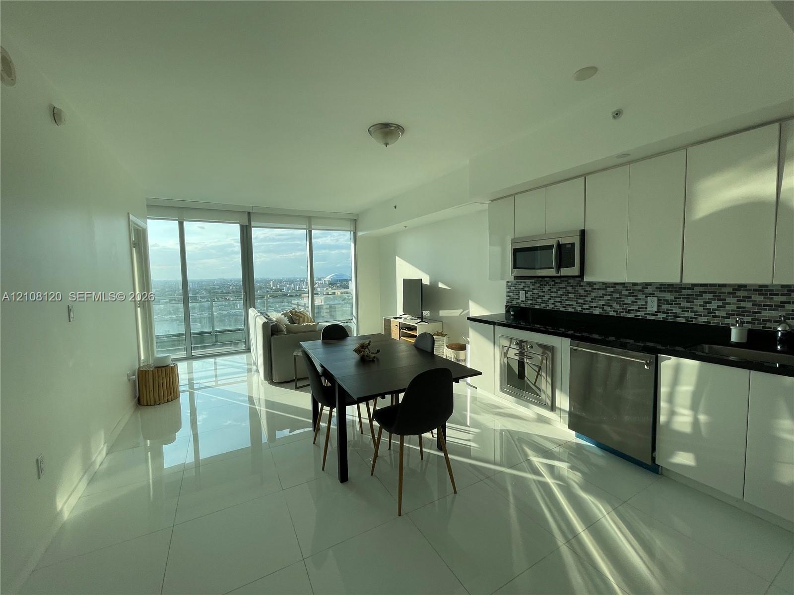Open-concept floor plan featuring high-gloss tile flooring, floor-to-ceiling glass paneling, white flat-panel cabinetry, stainless steel appliances, and a glass-tile mosaic backsplash