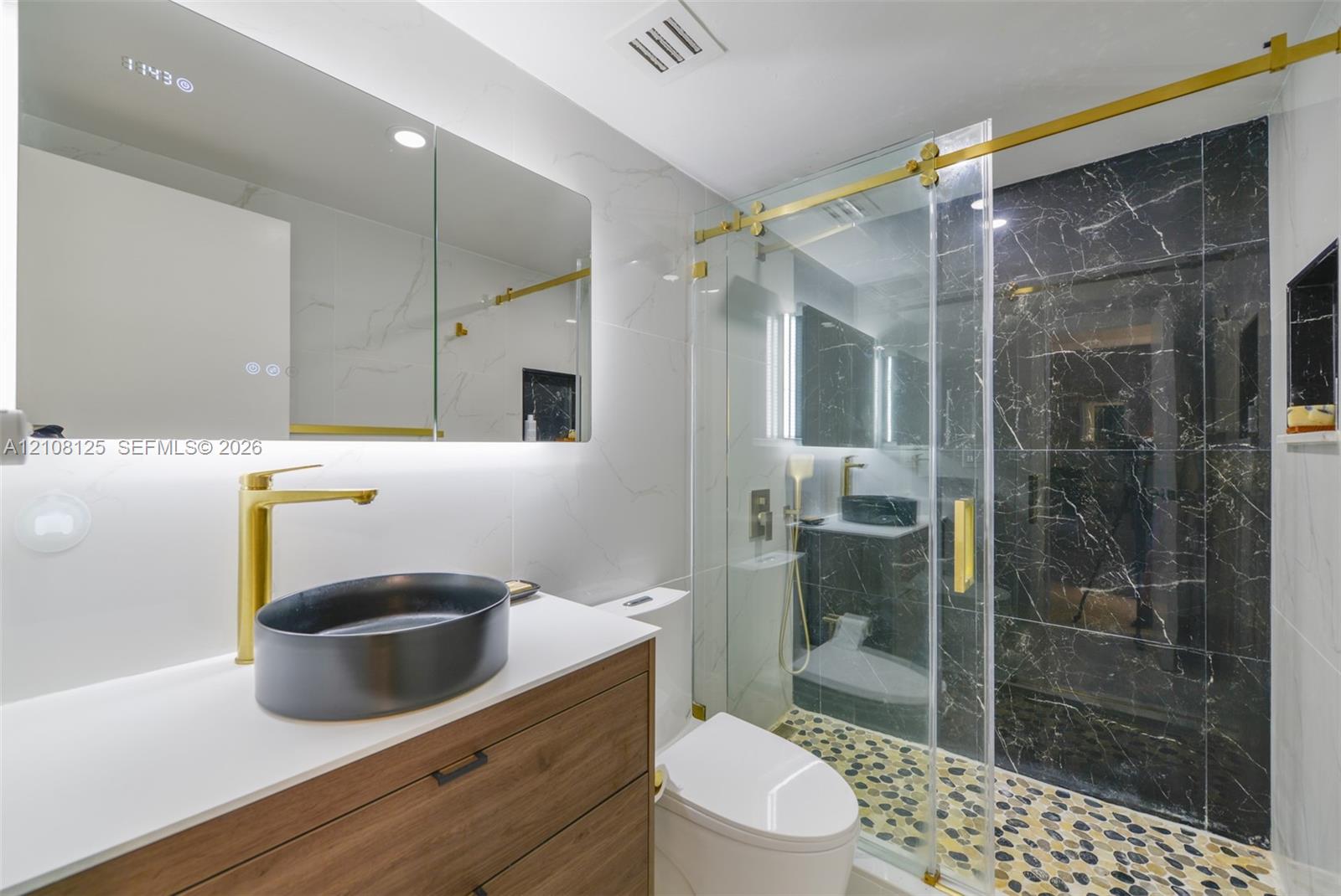 Bathroom featuring a wood-finish vanity with a quartz countertop, oval vessel sink, gold-toned hardware, and a walk-in shower with stone-tile flooring and dark accent tiling