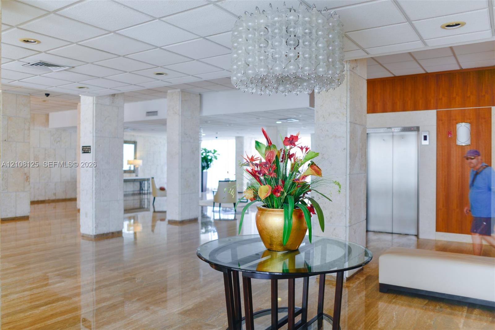 Spacious lobby featuring polished stone flooring, stone-clad structural columns, stainless steel elevator doors, and a decorative tiered pendant light fixture